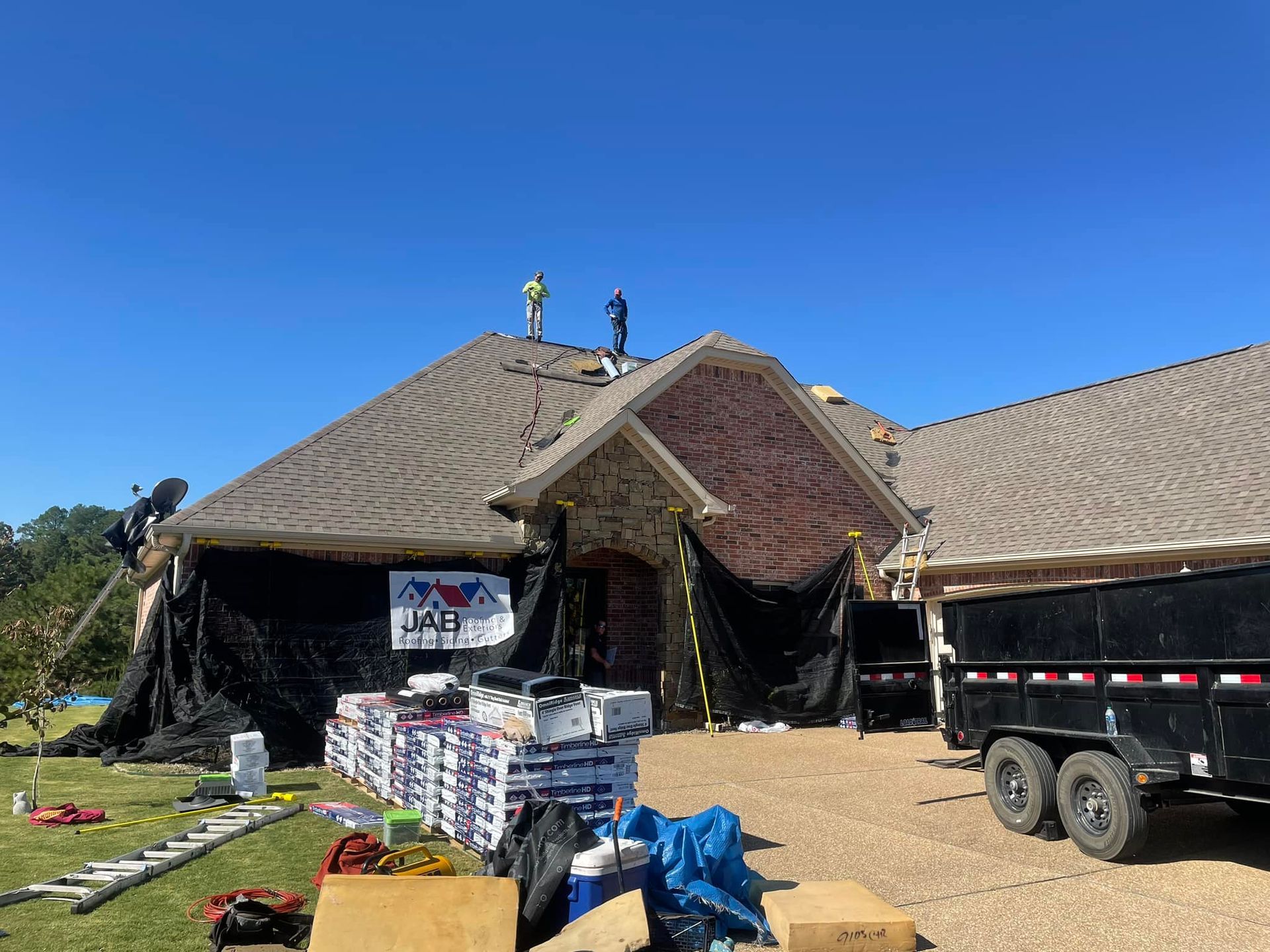 House with damaged roof and storm debris, pallets and truck in front under a clear blue sky