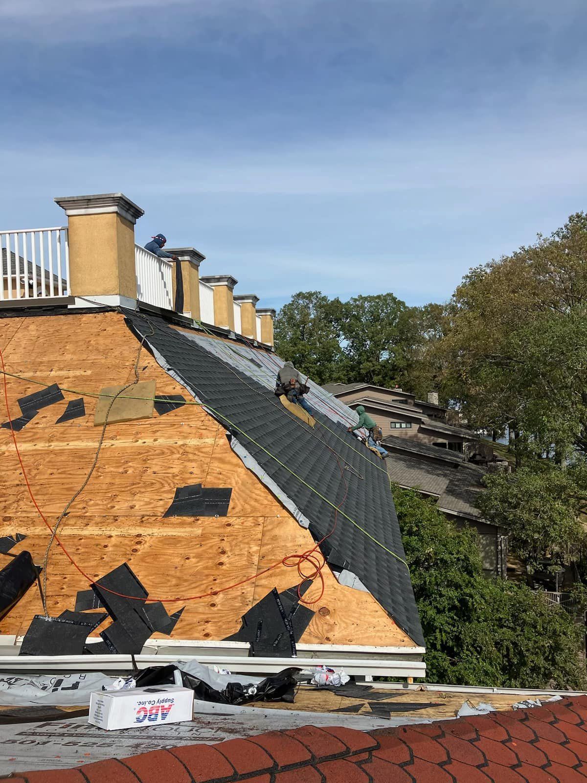 Damaged house roof with exposed plywood and tarp, chimney stacks, and fallen debris beside trees.