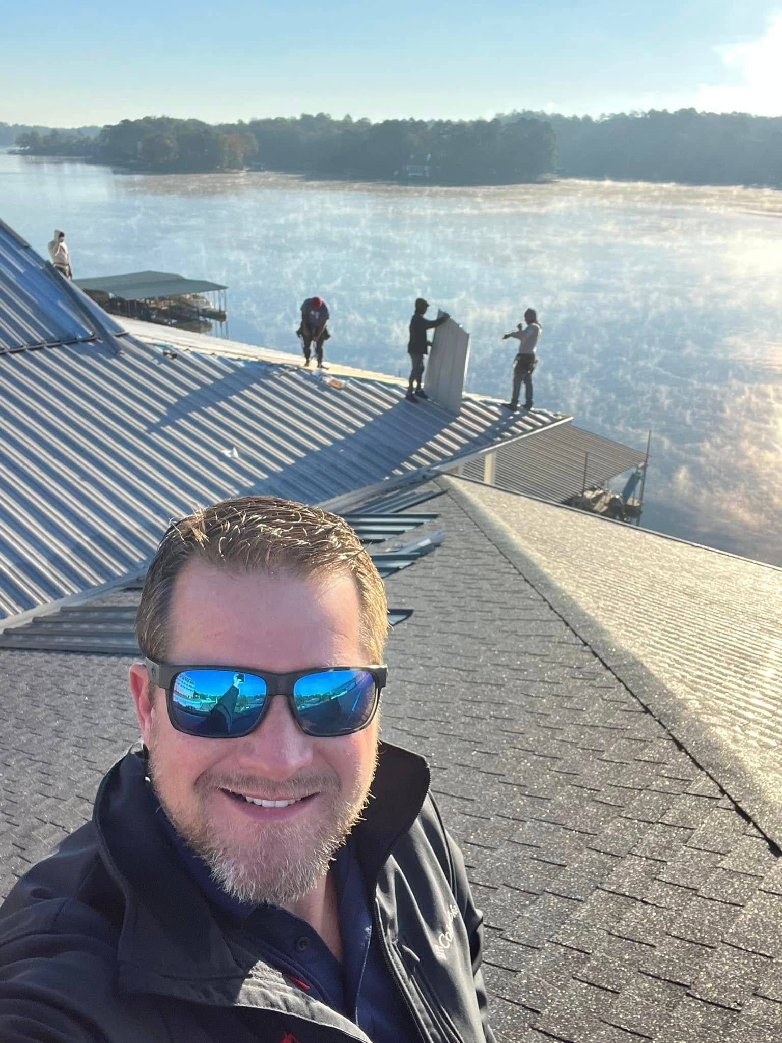 Man smiling in sunglasses on a rooftop overlooking a bright waterfront with several people near the edge.