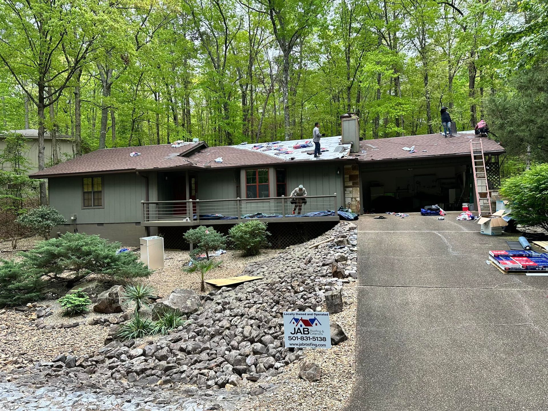 House with a long driveway and rock landscaping in a wooded setting, with a few people on the roof.