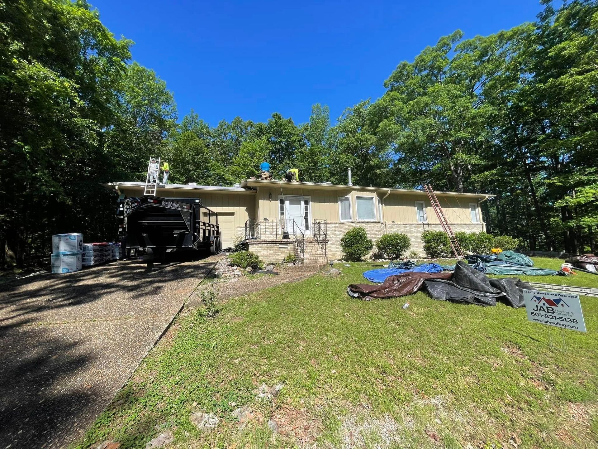 Single-story house with a gravel driveway and scattered materials in a wooded yard under a blue sky
