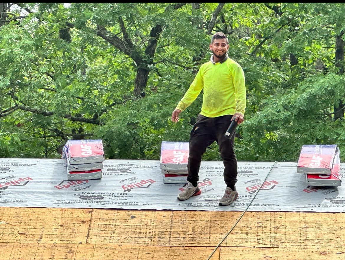 Person in a neon yellow jacket standing on a rooftop in front of trees and three small boxes.