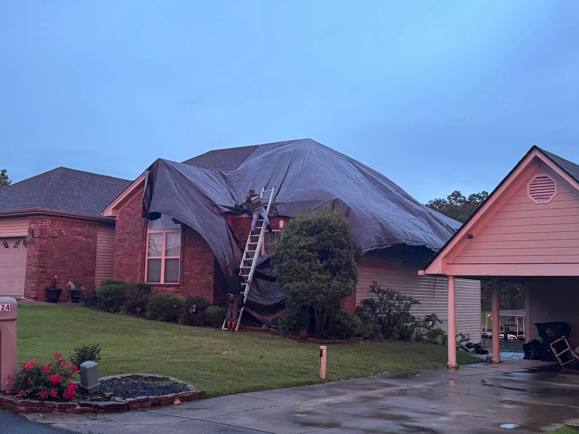 Brick house with a collapsed roof tarp, ladder, and driveway carport during storm damage recovery