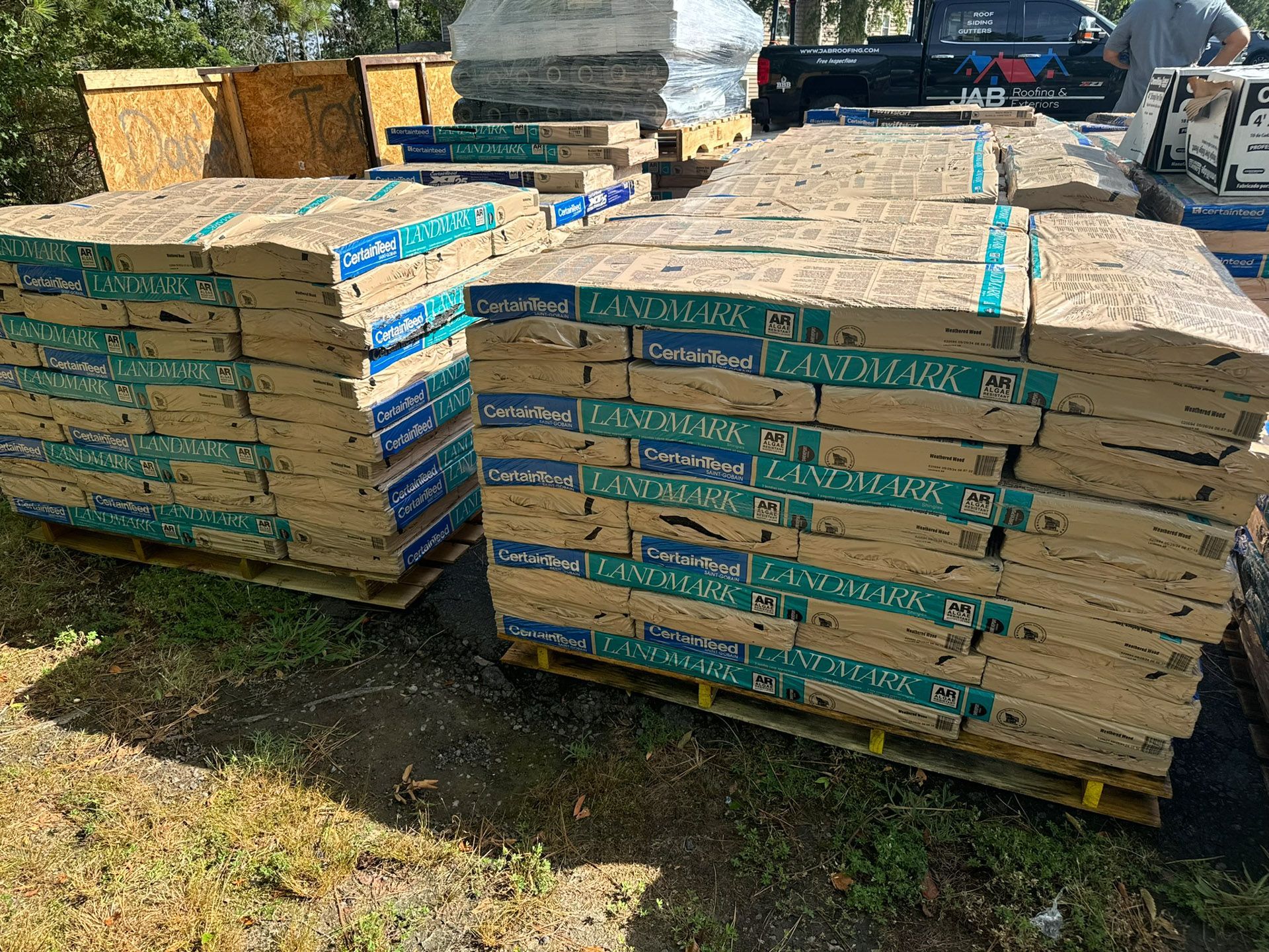 Stacks of lumber wrapped in teal straps on pallets in an outdoor yard, with a worker in the background