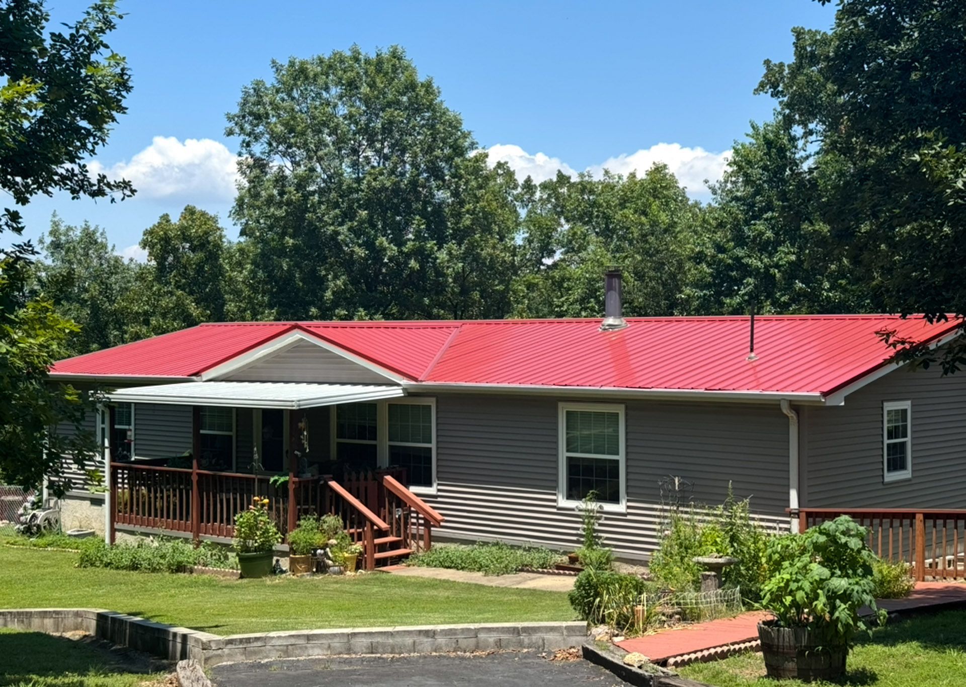 Single-story gray house with a red roof and front porch in a sunny, landscaped yard