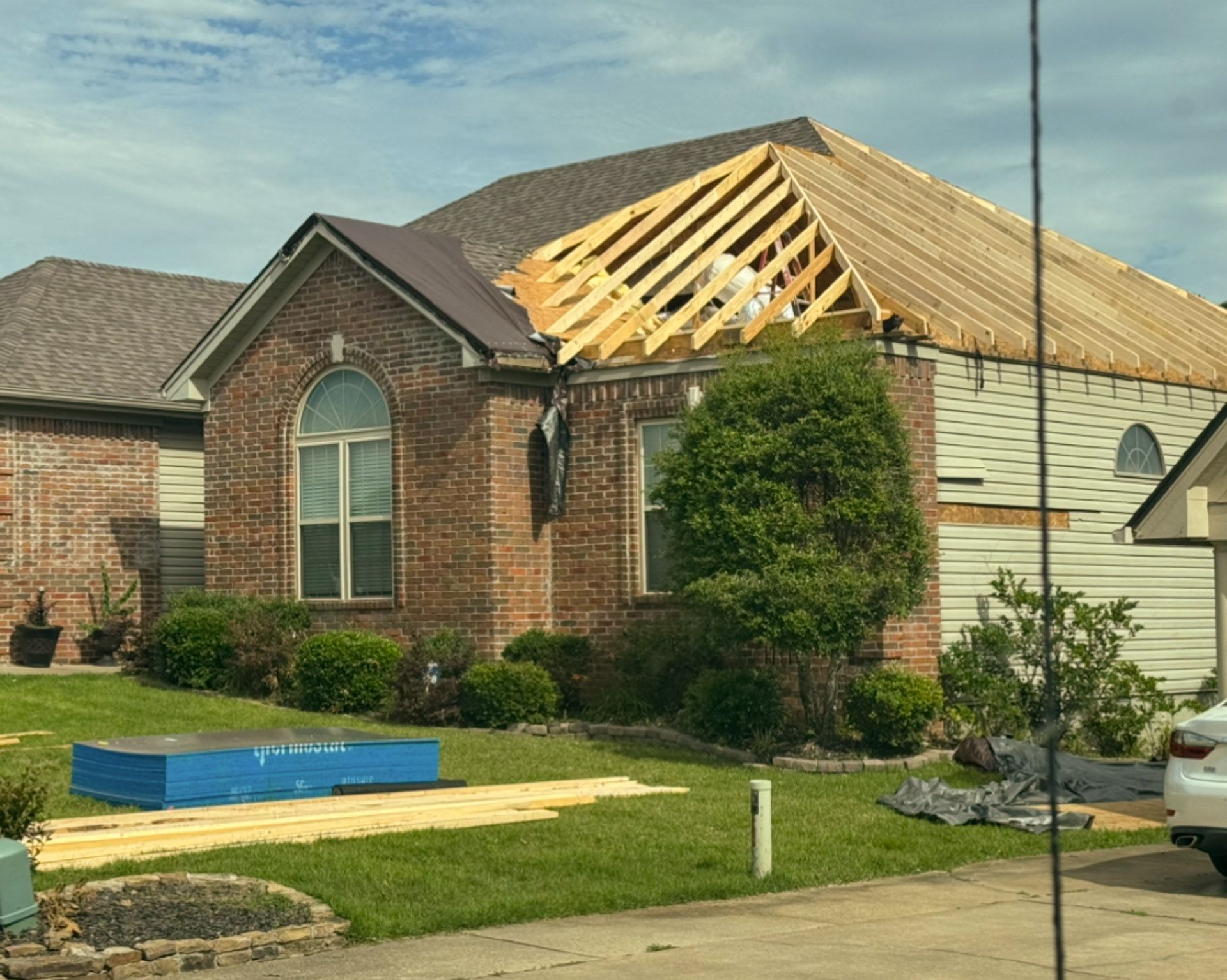 Brick house with roof under construction, exposed wood framing, and a blue dumpster in the front yard.
