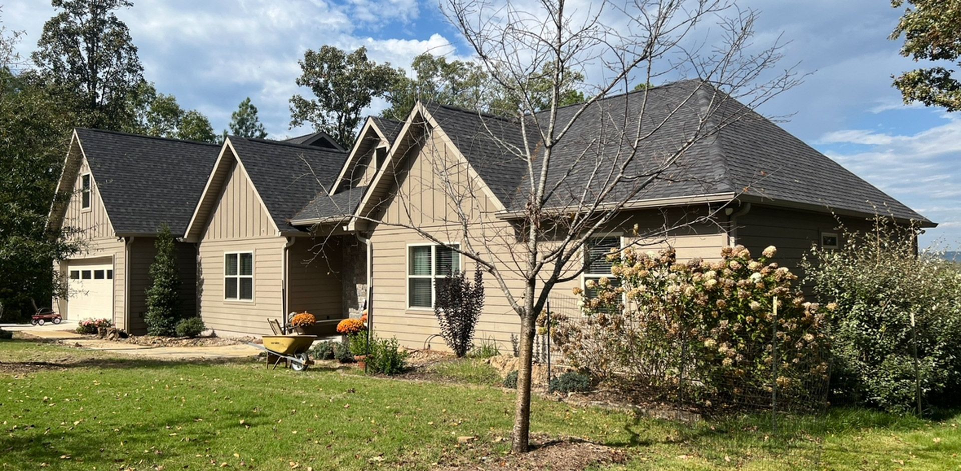 Two beige houses with dark roofs in a green yard, surrounded by trees and shrubs