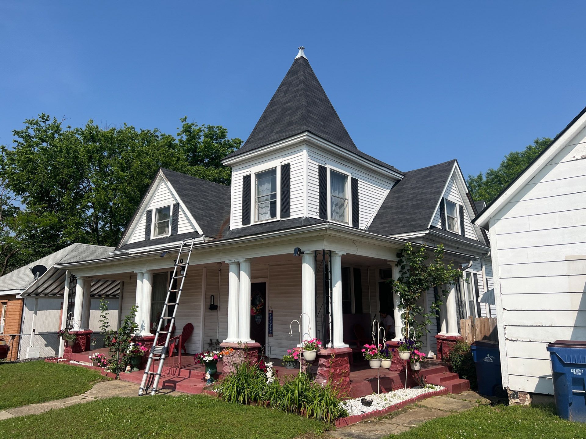 Victorian house with a steep roof, white porch columns, and flower beds under a clear blue sky