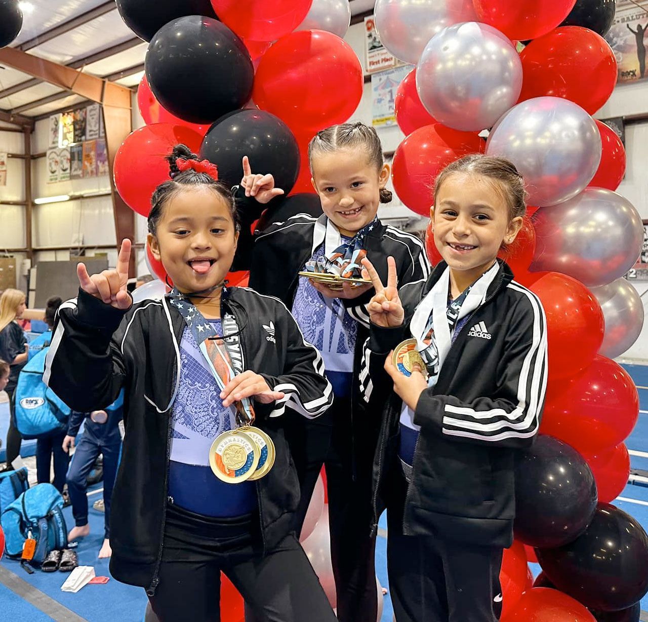 Three children in matching athletic gear pose with medals in front of a red, silver, and black balloon arch at a gym.