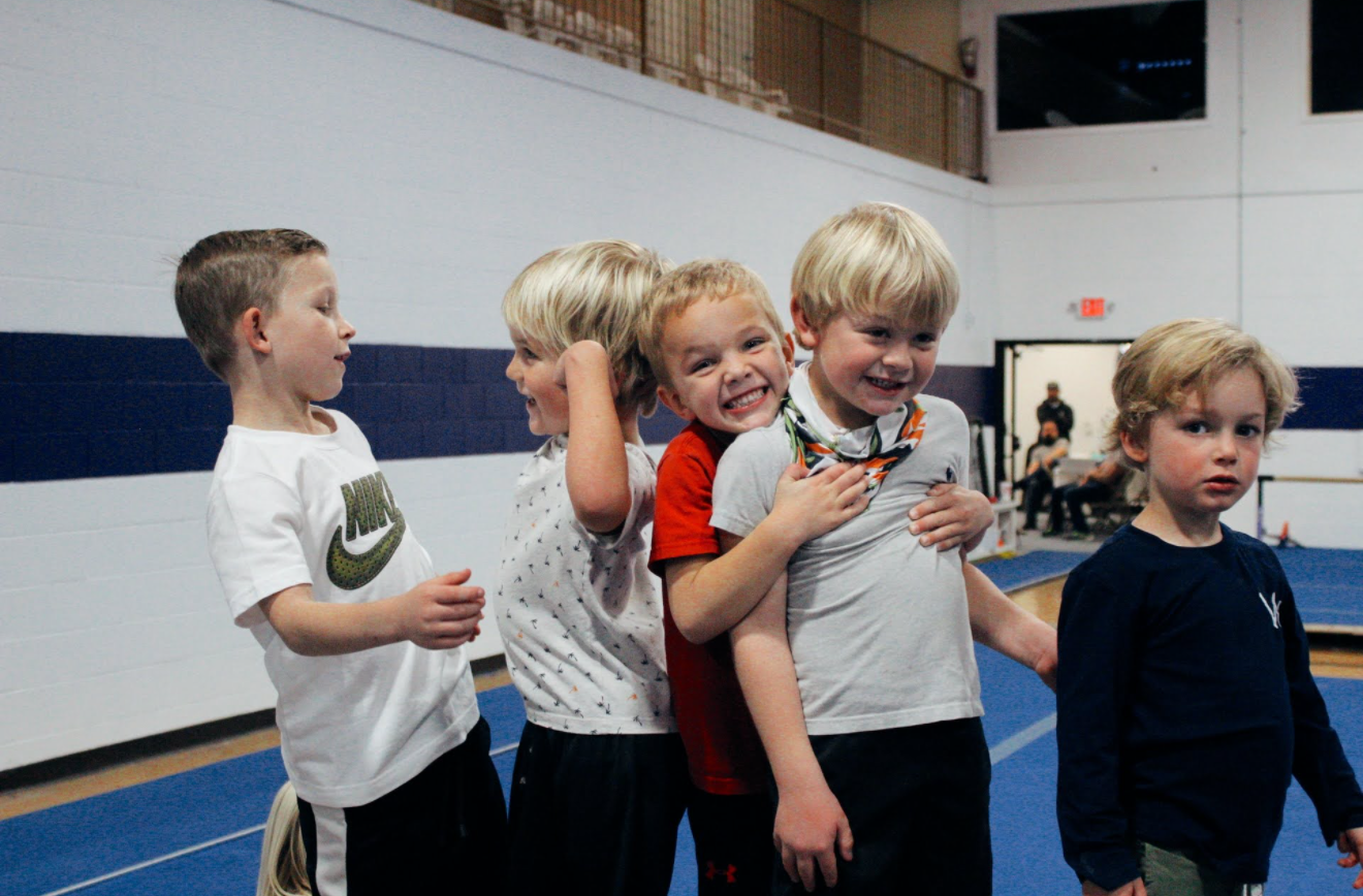 Five children line up on a blue gymnastics mat; four are huddled together smiling while one looks forward.