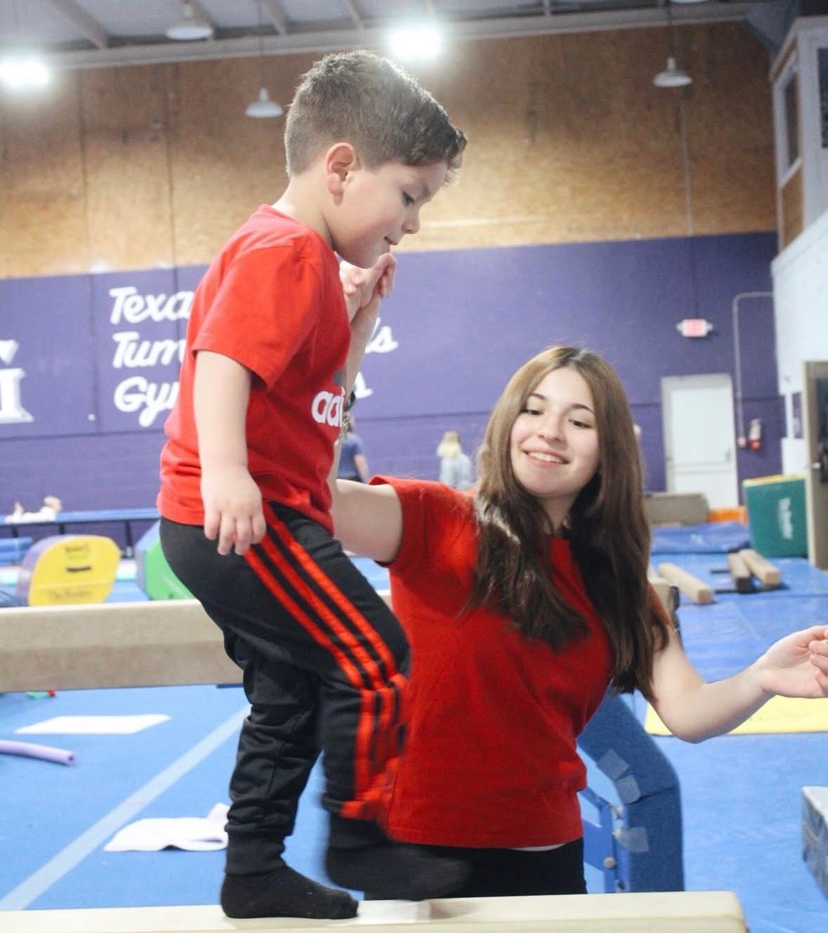 A gymnastics coach in a red shirt helps a child in a red shirt and black sweatpants balance on a gymnastics beam.