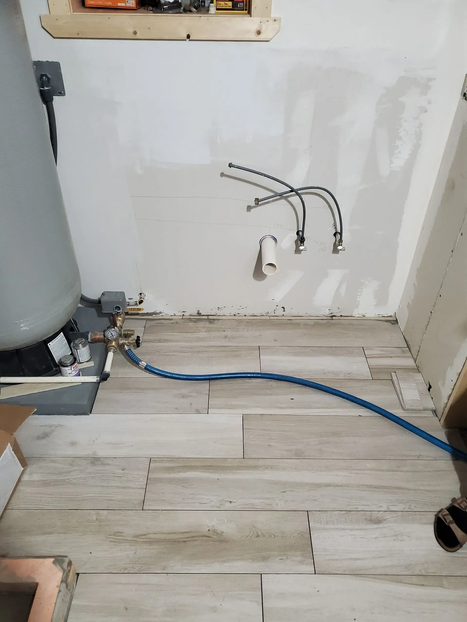 Utility room with exposed plumbing, wall hoses, and a blue cable on a light wood floor.