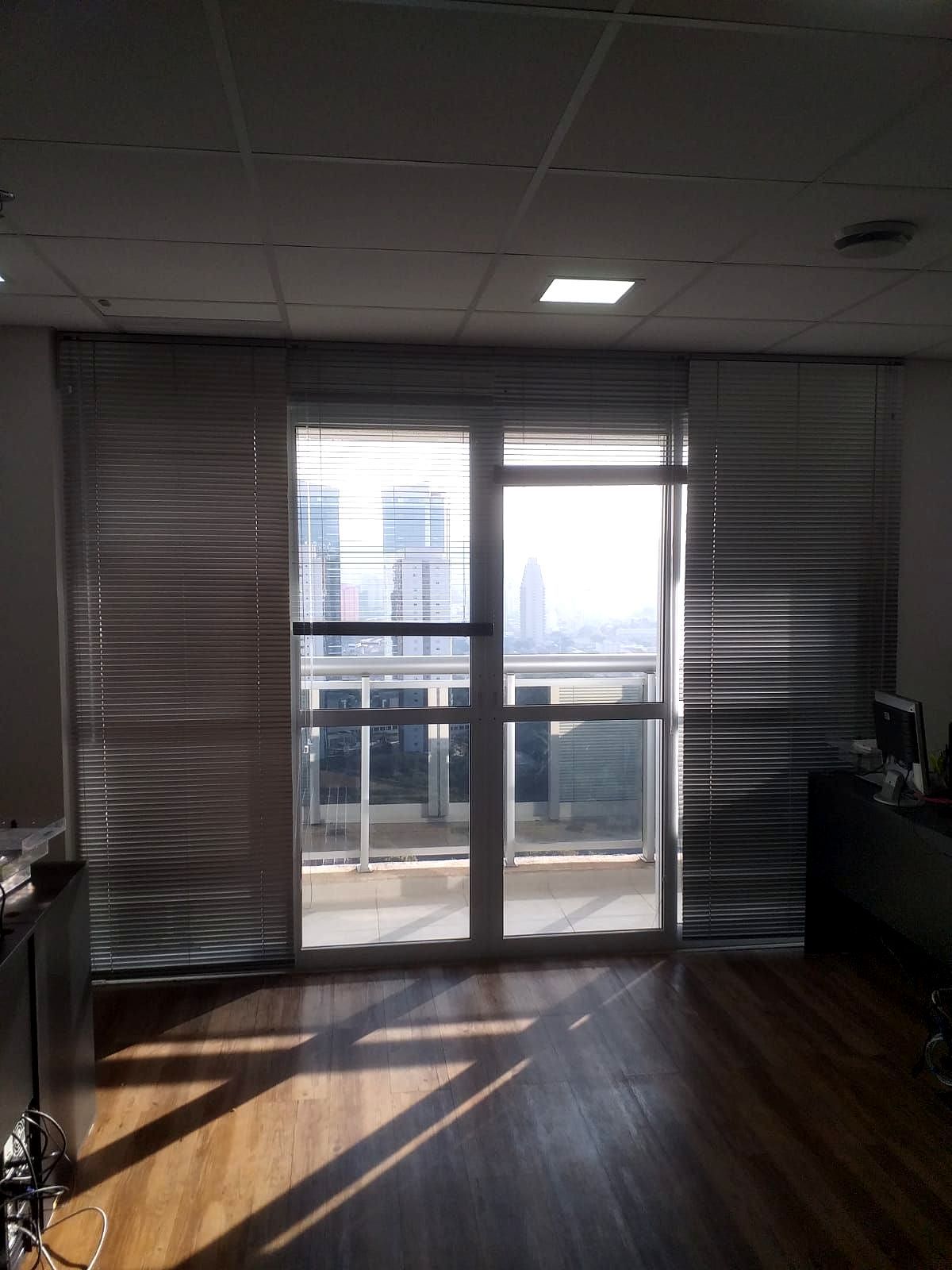 Escritório pouco iluminado com grandes janelas cobertas por persianas nejflex de alumínio, com vista para a paisagem urbana lá fora.
