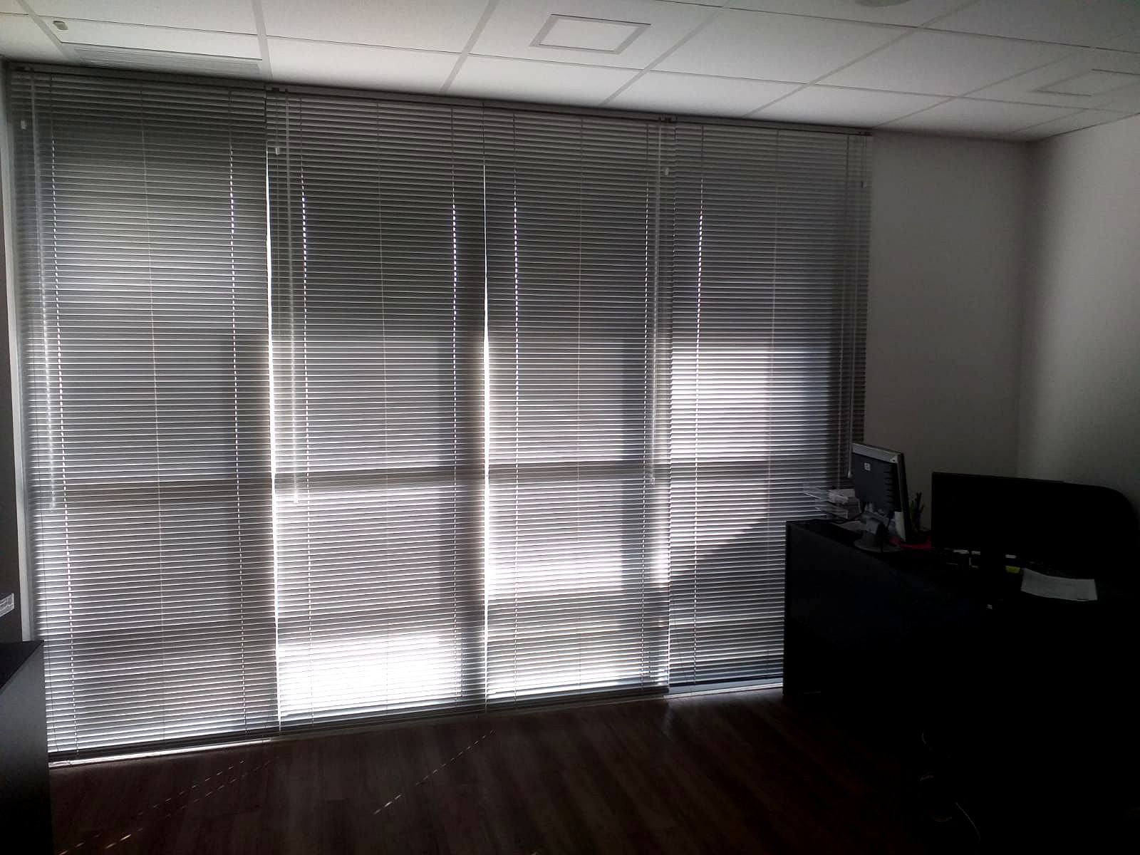 Interior de escritório com persianas horizontais de alumínio fechadas filtrando a luz solar, uma mesa escura à direita e piso de madeira.