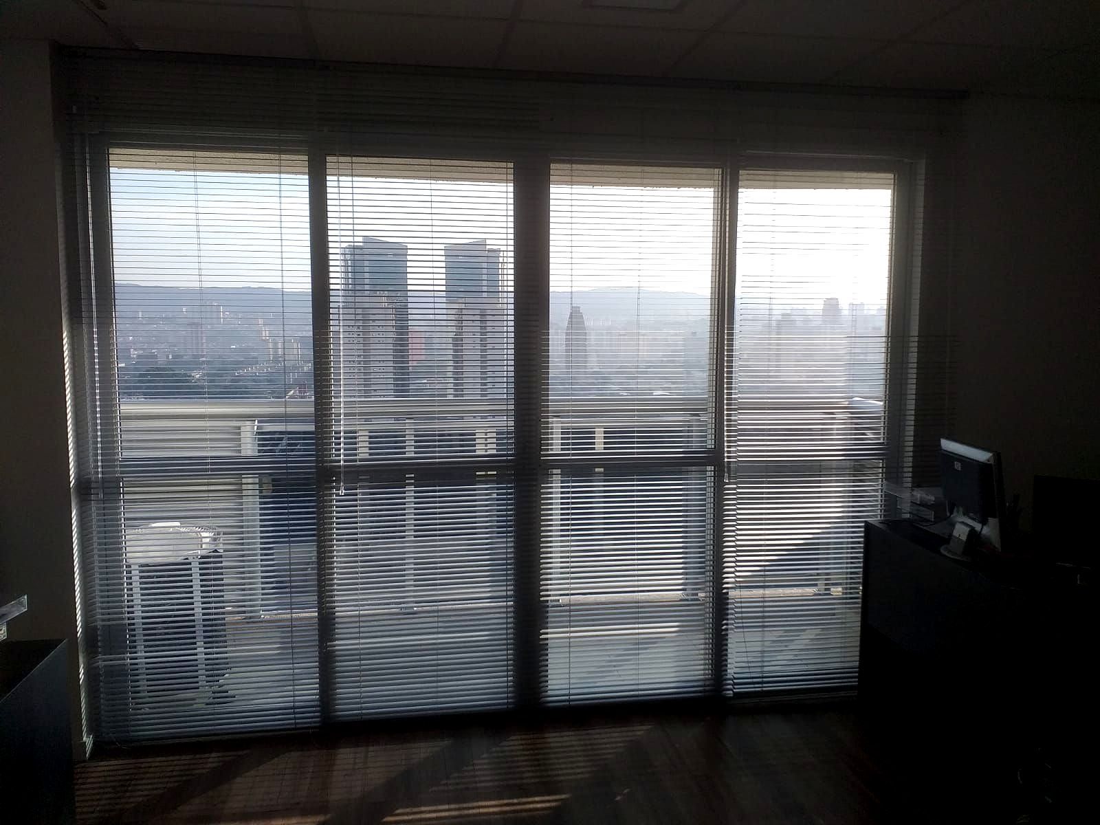 Escritório pouco iluminado com grandes janelas cobertas por persianas nejflex de alumínio, com vista para a paisagem urbana lá fora.