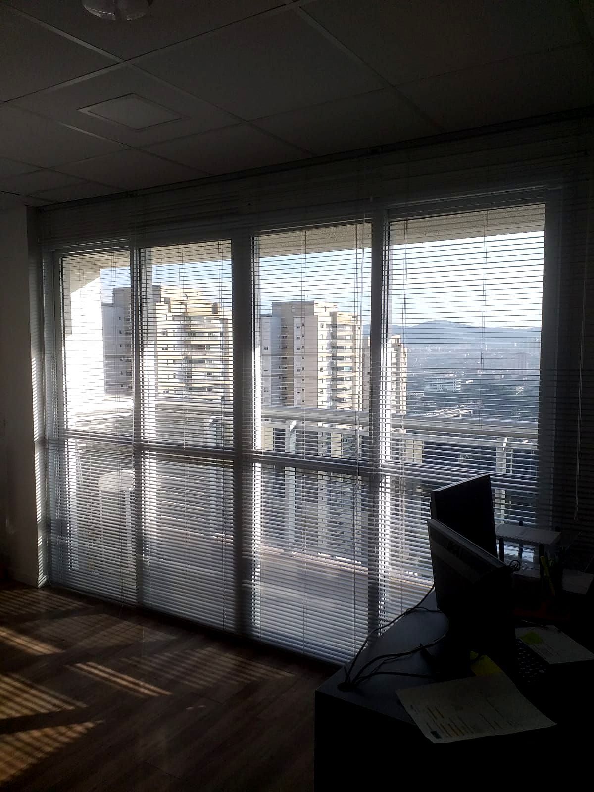 Escritório pouco iluminado com grandes janelas cobertas por persianas nejflex de alumínio, com vista para a paisagem urbana lá fora.