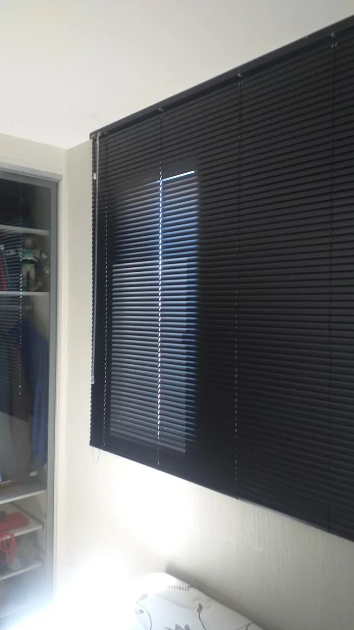 Persianas nejflex de alumínio pretas cobrindo uma janela, com a luz do sol penetrando pelas frestas estreitas.