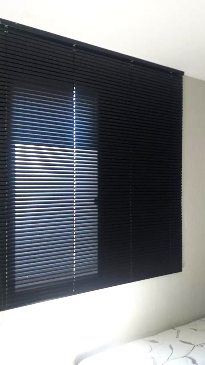 Persianas de alumínio pretas parcialmente fechadas ao lado de uma cama em um quarto de paredes brancas.