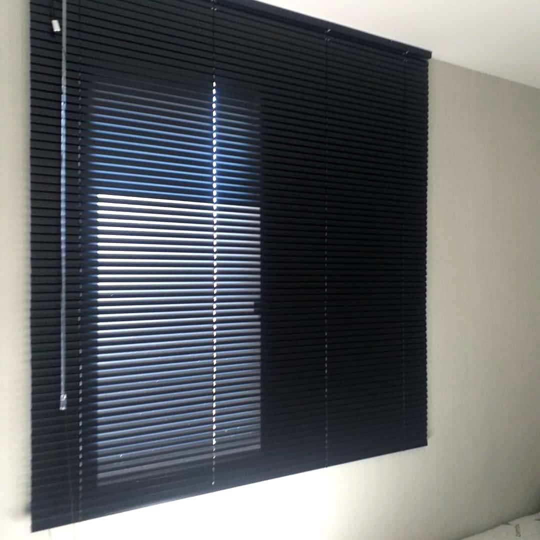 Persianas horizontais de alumínio pretas parcialmente fechadas sobre uma janela iluminada em um quarto de cores claras.