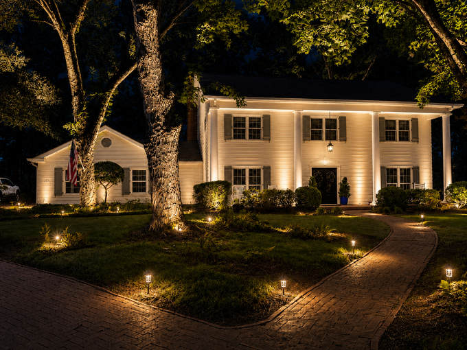 Illuminated two-story white house with lit windows, landscaped lawn, and trees at night
