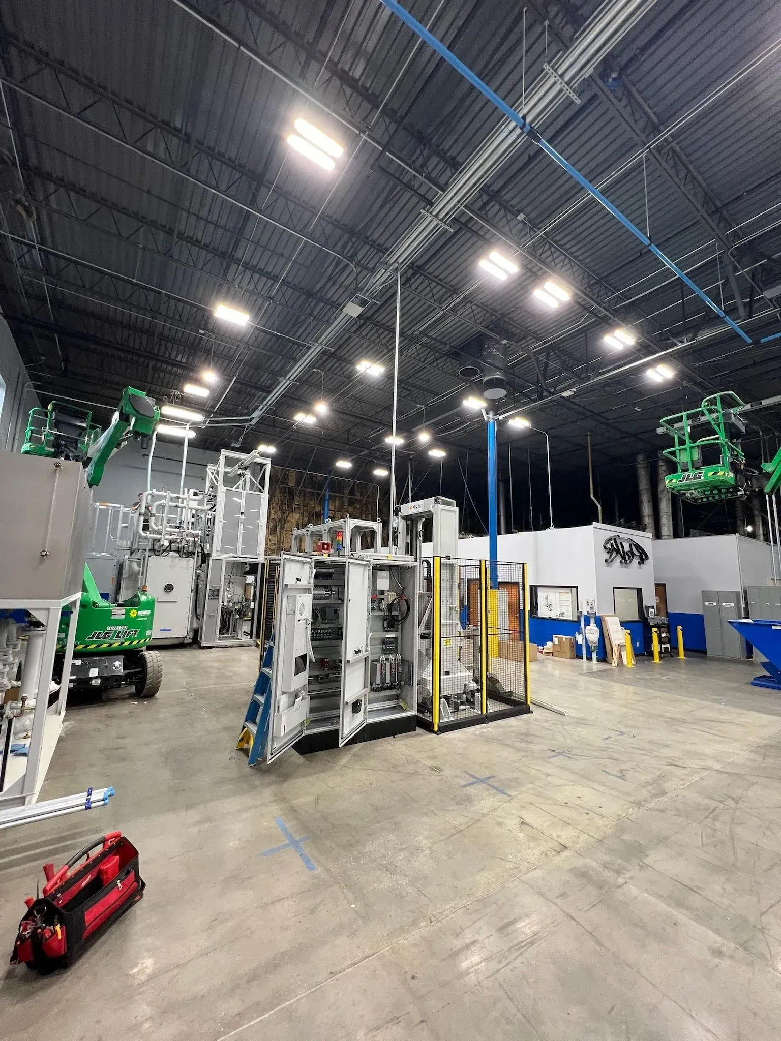 Large warehouse with industrial machines, overhead lights, and a concrete floor, with a small red cart in front