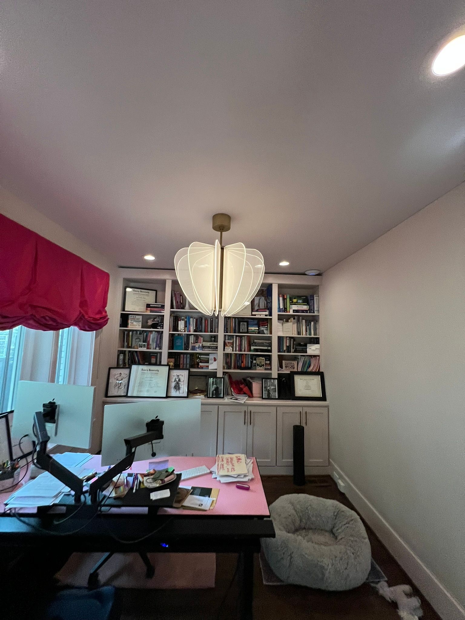 Home office with desk, shelves, white pendant light, pink curtain, and a gray beanbag by the wall.
