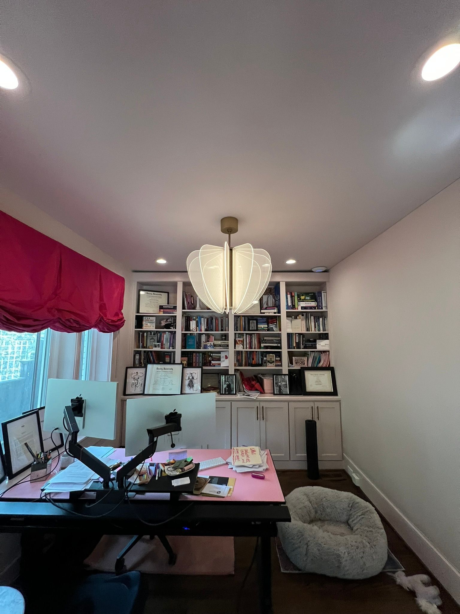 Home office with desk, chair, bookshelves, white walls, and a large hanging pendant light.
