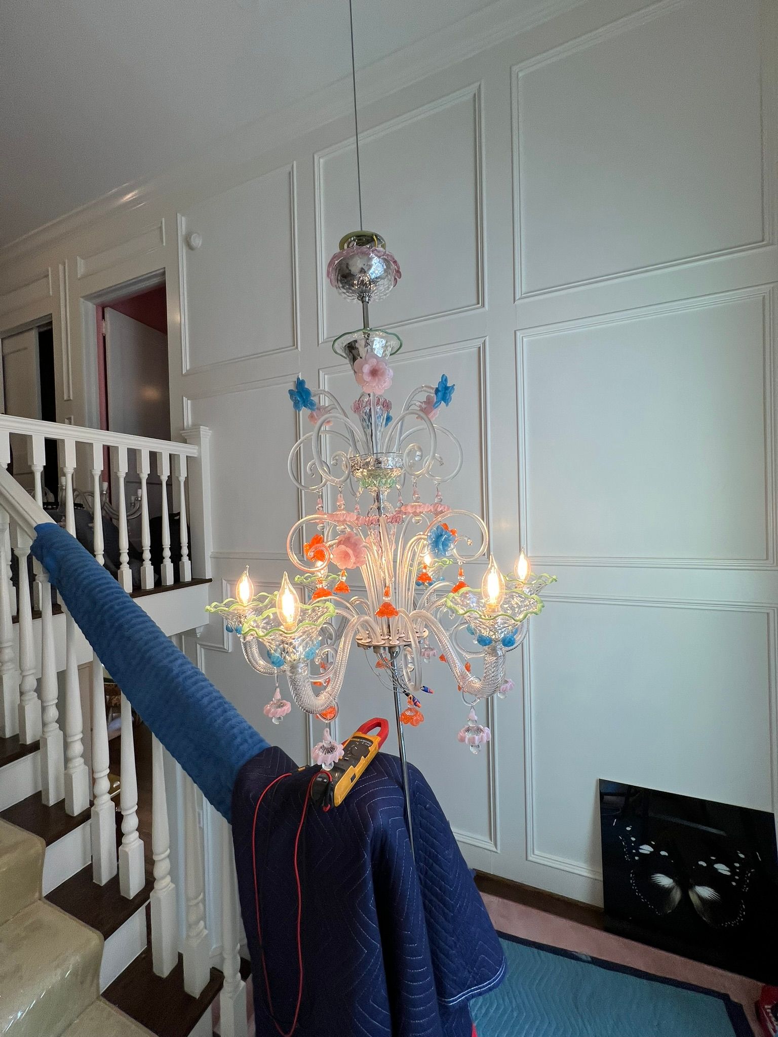 Decorative chandelier with colorful hanging ornaments in a white-paneled hallway over a stair railing