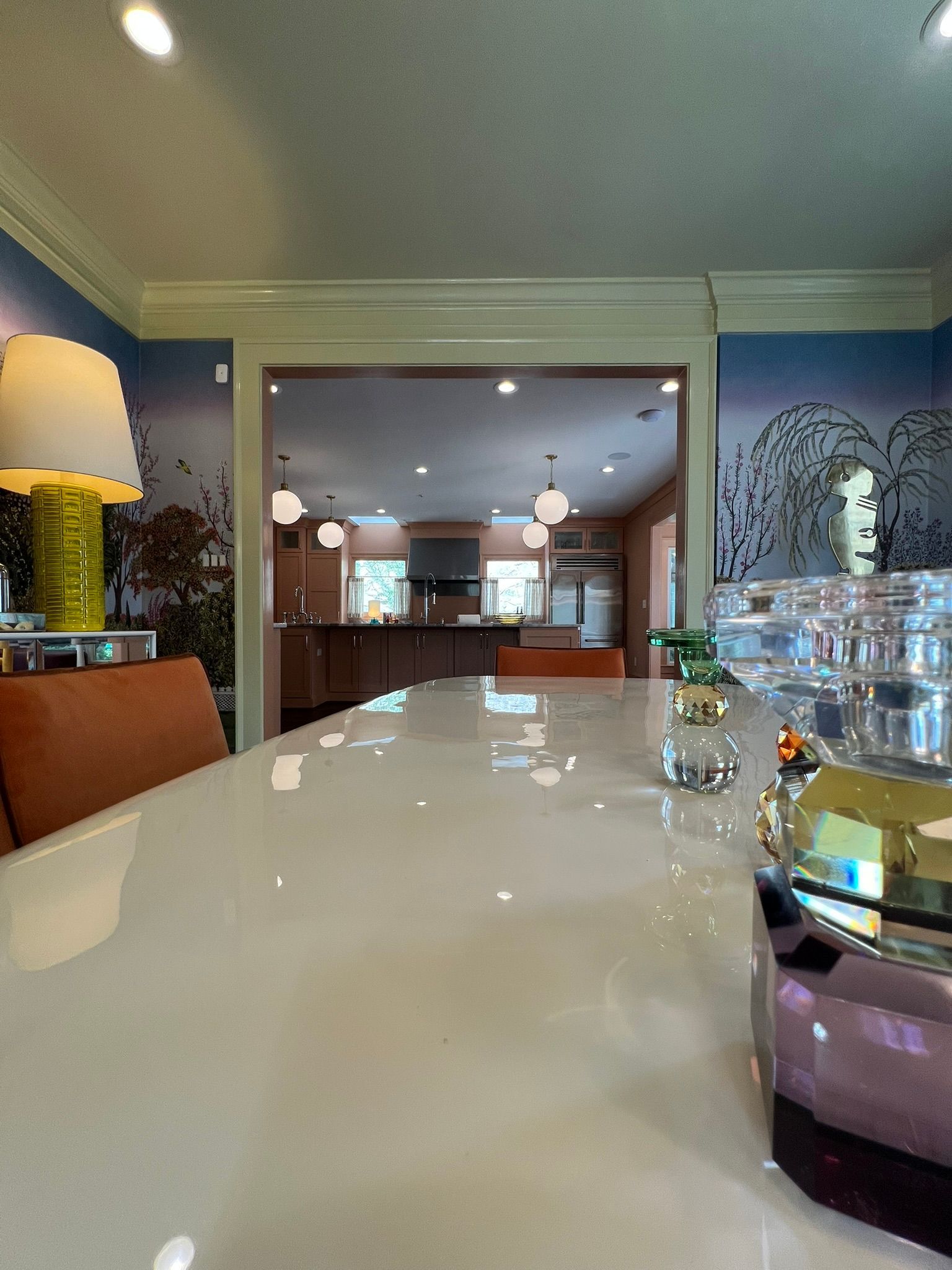 Dining room with a glossy white table, warm lamp, and open view into a kitchen under recessed lights.