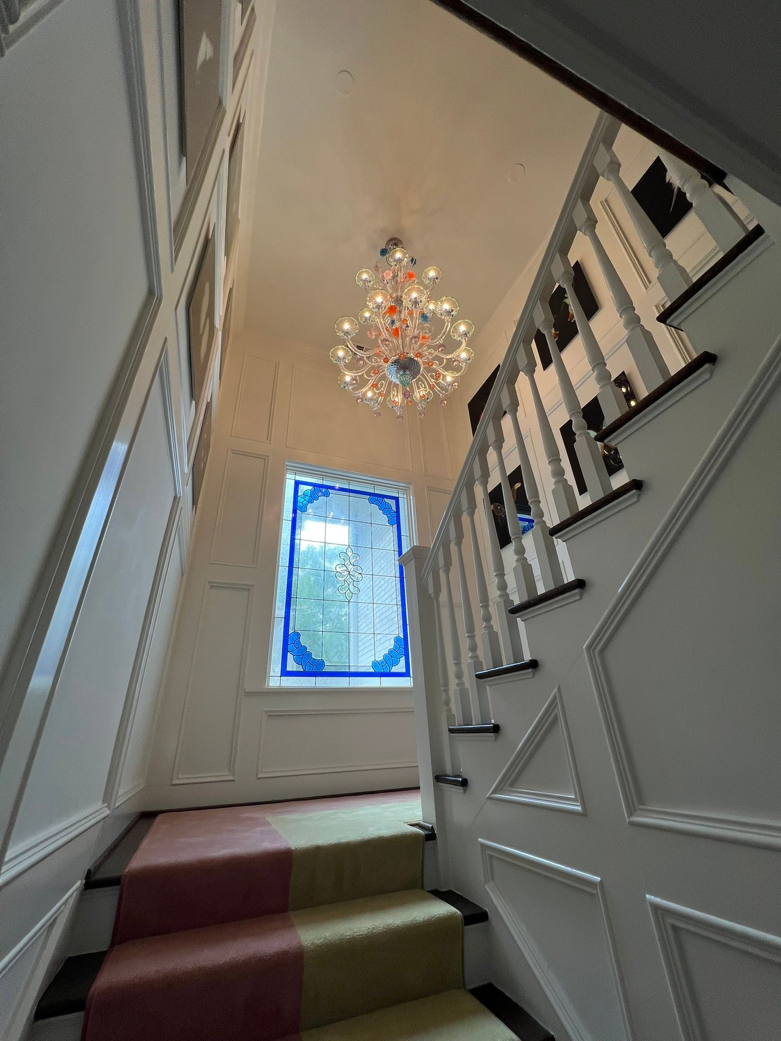 Narrow staircase with white paneling, patterned runner, and a chandelier beneath a stained-glass window.