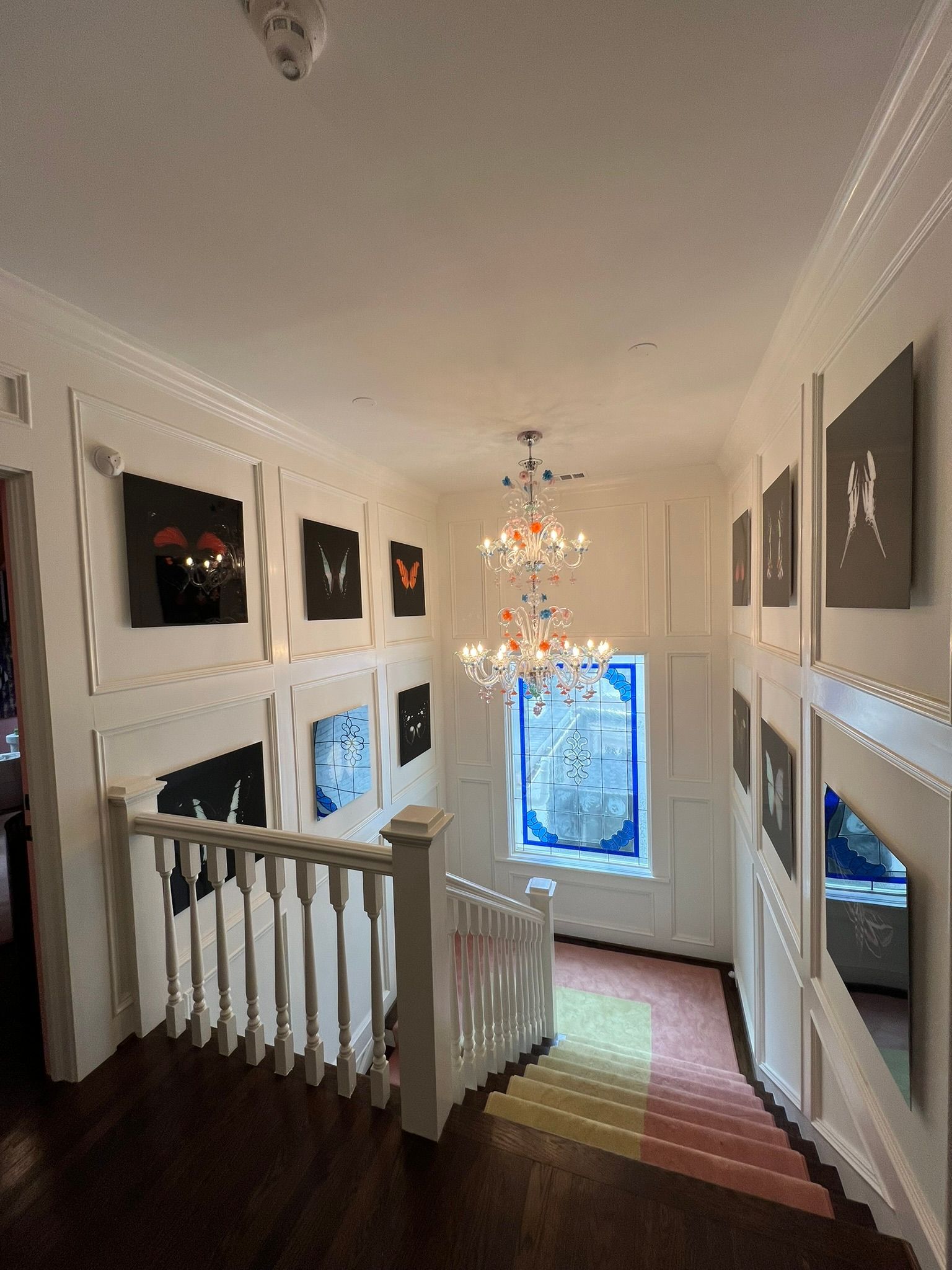 Bright hallway with framed art, a staircase, a hanging chandelier, and a window at the far end.