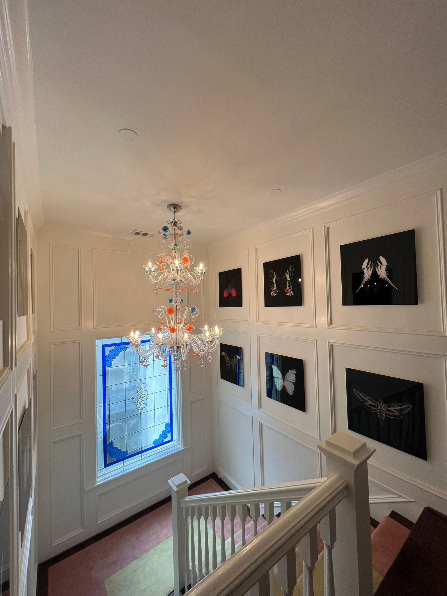 Bright stairwell with chandelier, white railings, framed black artworks, and a blue-lit window.