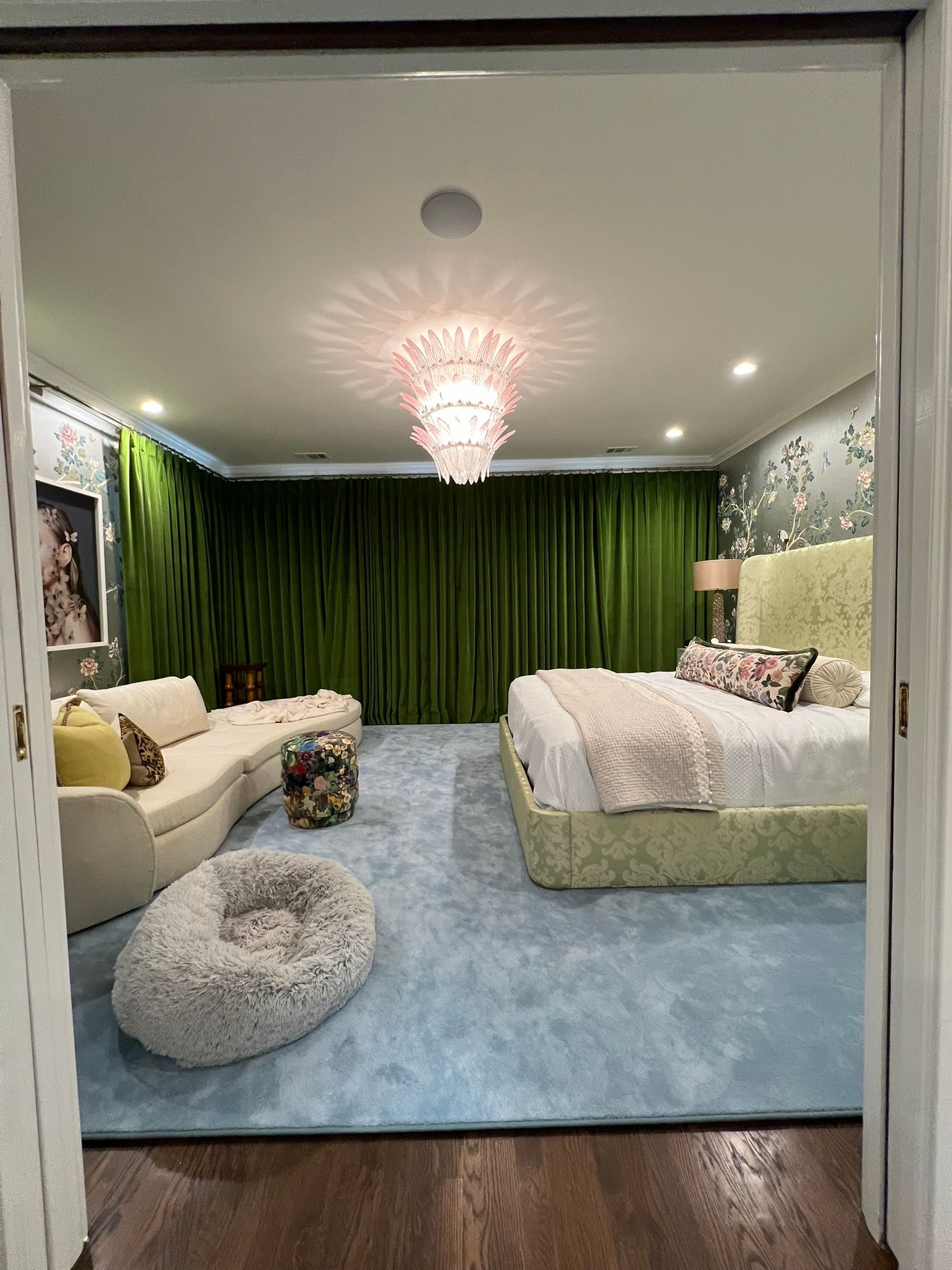 Modern bedroom with green curtains, blue rug, bed, sofa, and chandelier in soft lighting