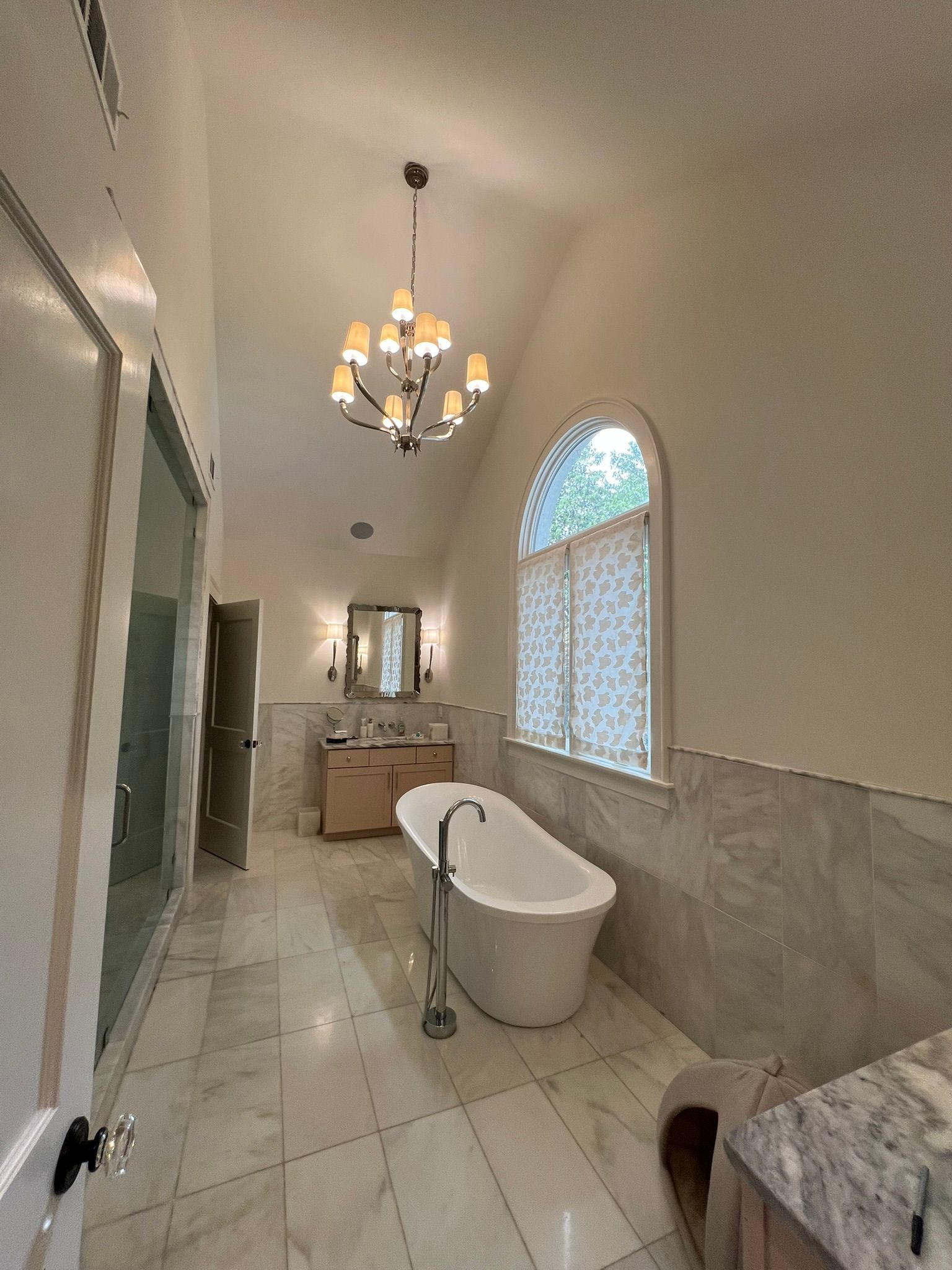 Spacious bathroom with a chandelier, freestanding tub, double vanity, and arched window with blinds