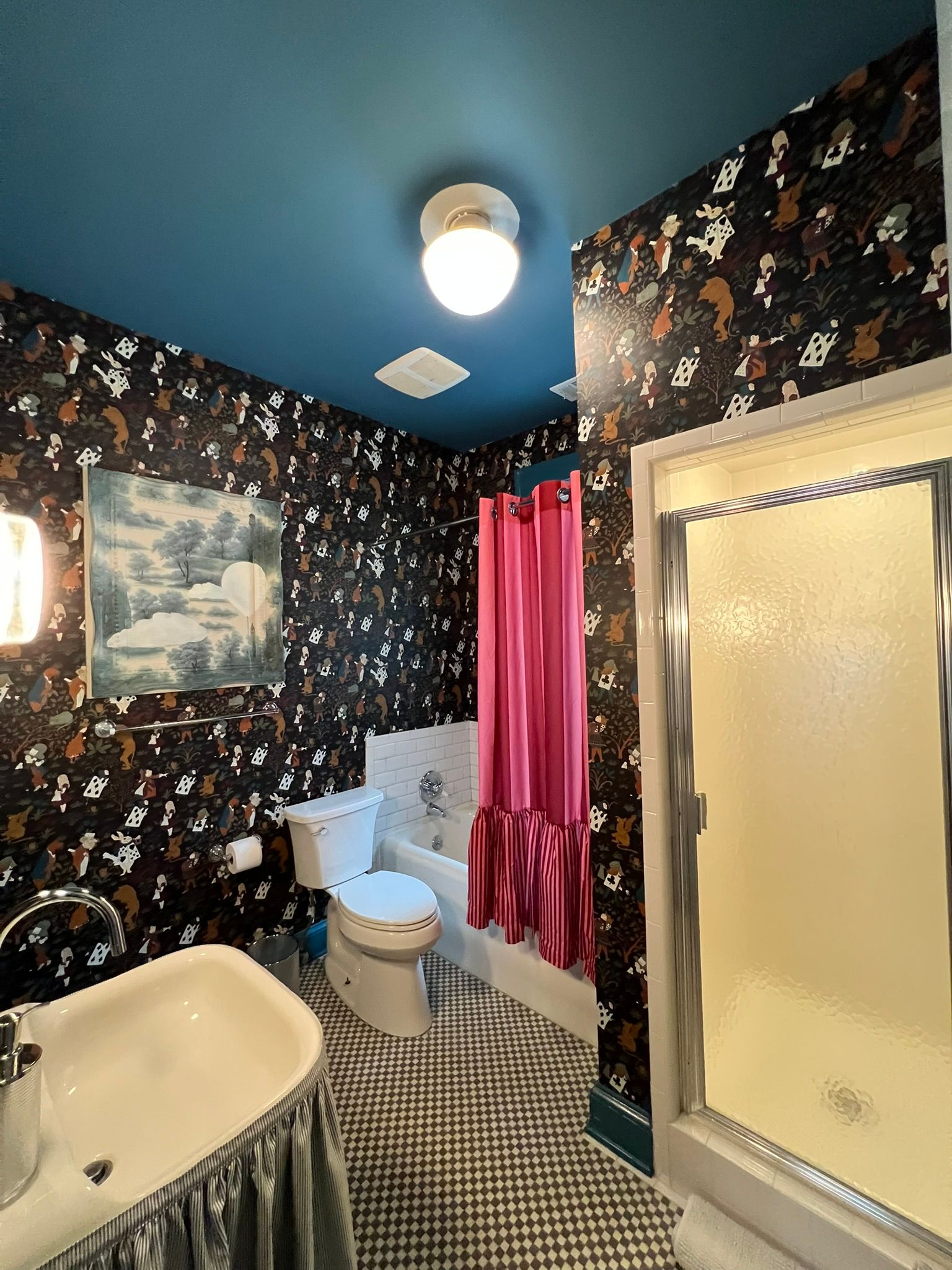 Small bathroom with floral wallpaper, pink shower curtain, frosted shower door, toilet, sink, and blue ceiling.