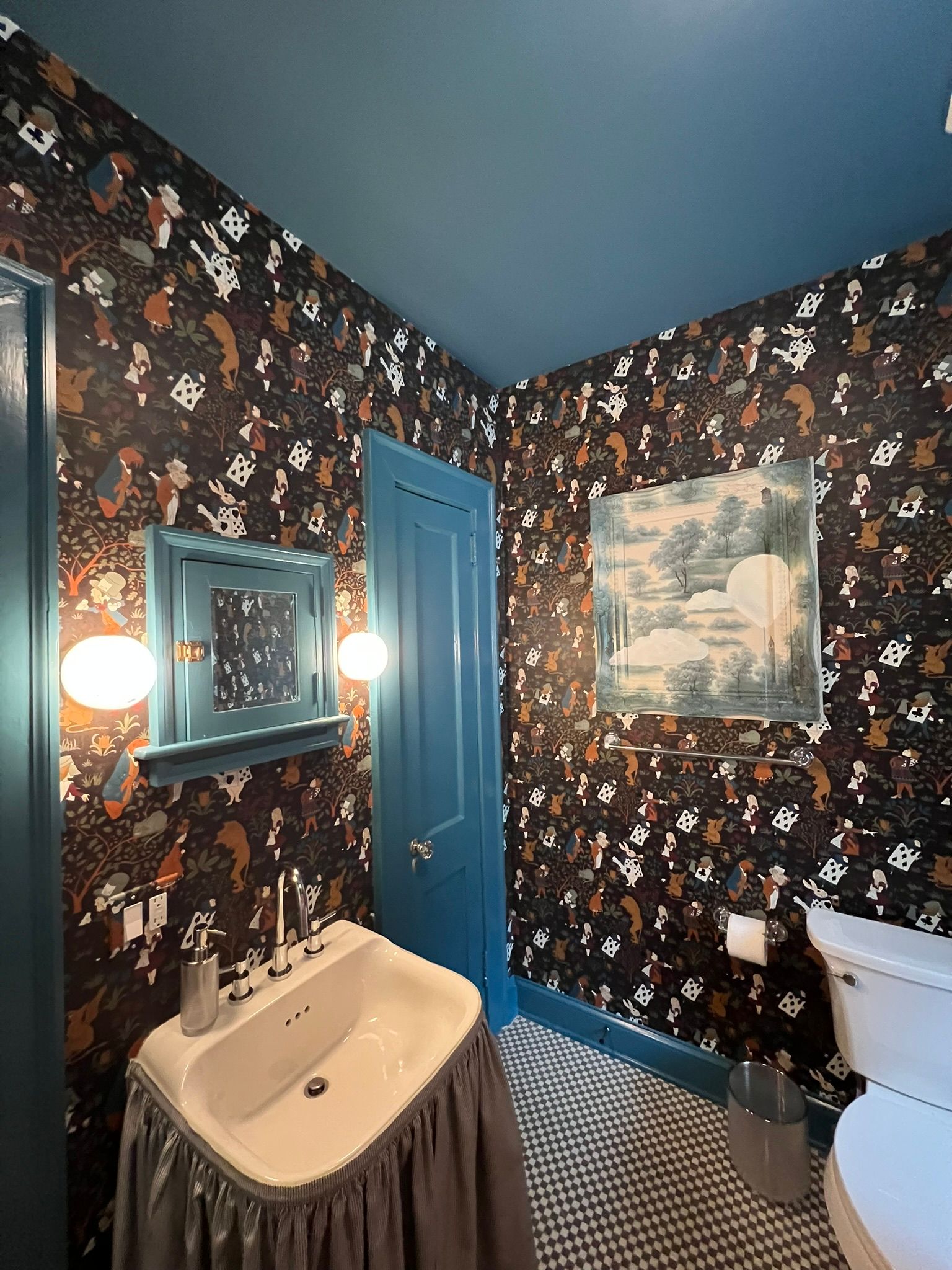 Compact bathroom with floral wallpaper, blue ceiling and door, pedestal sink, mirror, and toilet.