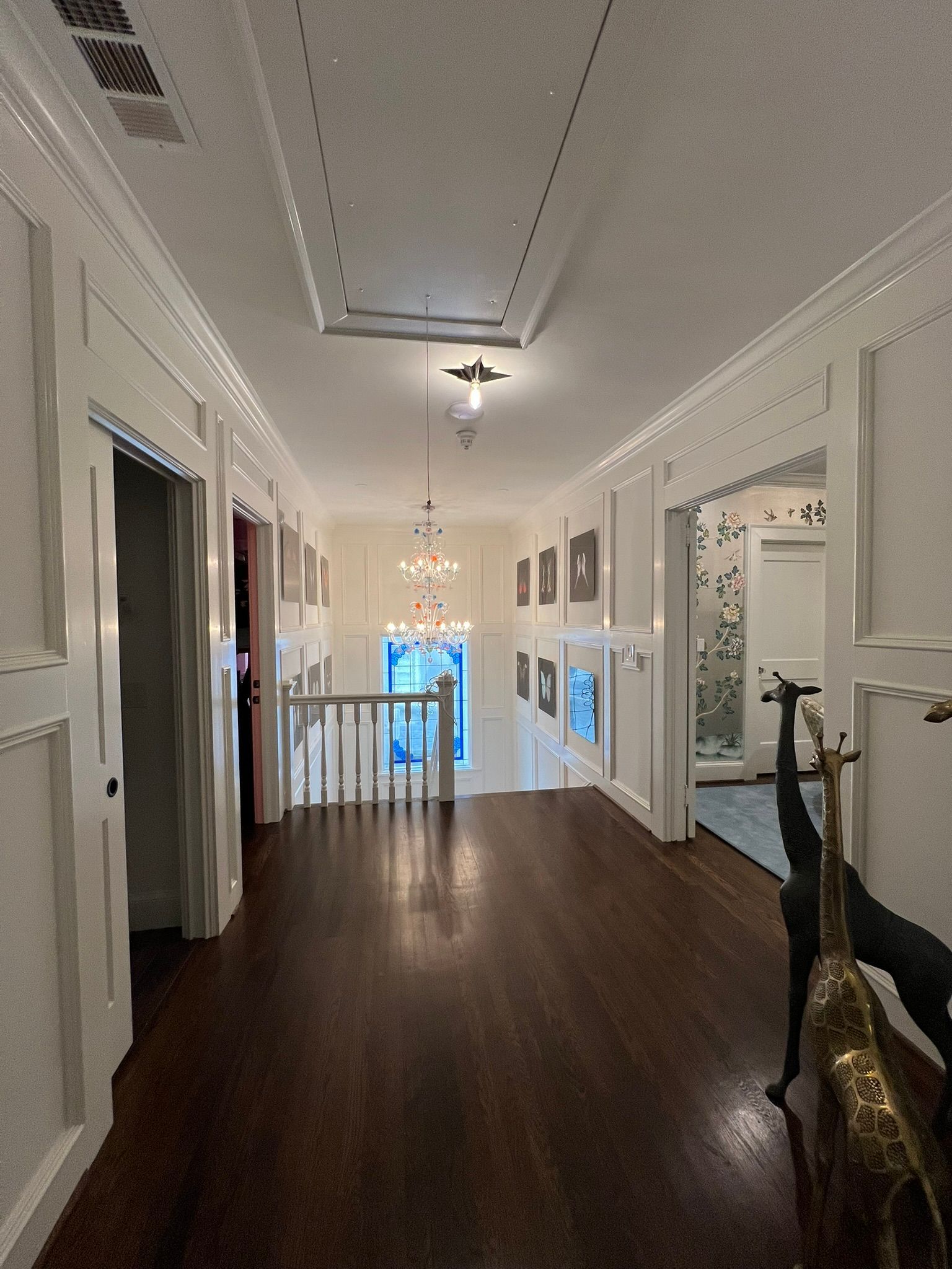 Elegant hallway with dark wood floors, white walls, chandeliers, and a view to the lower level through a railing