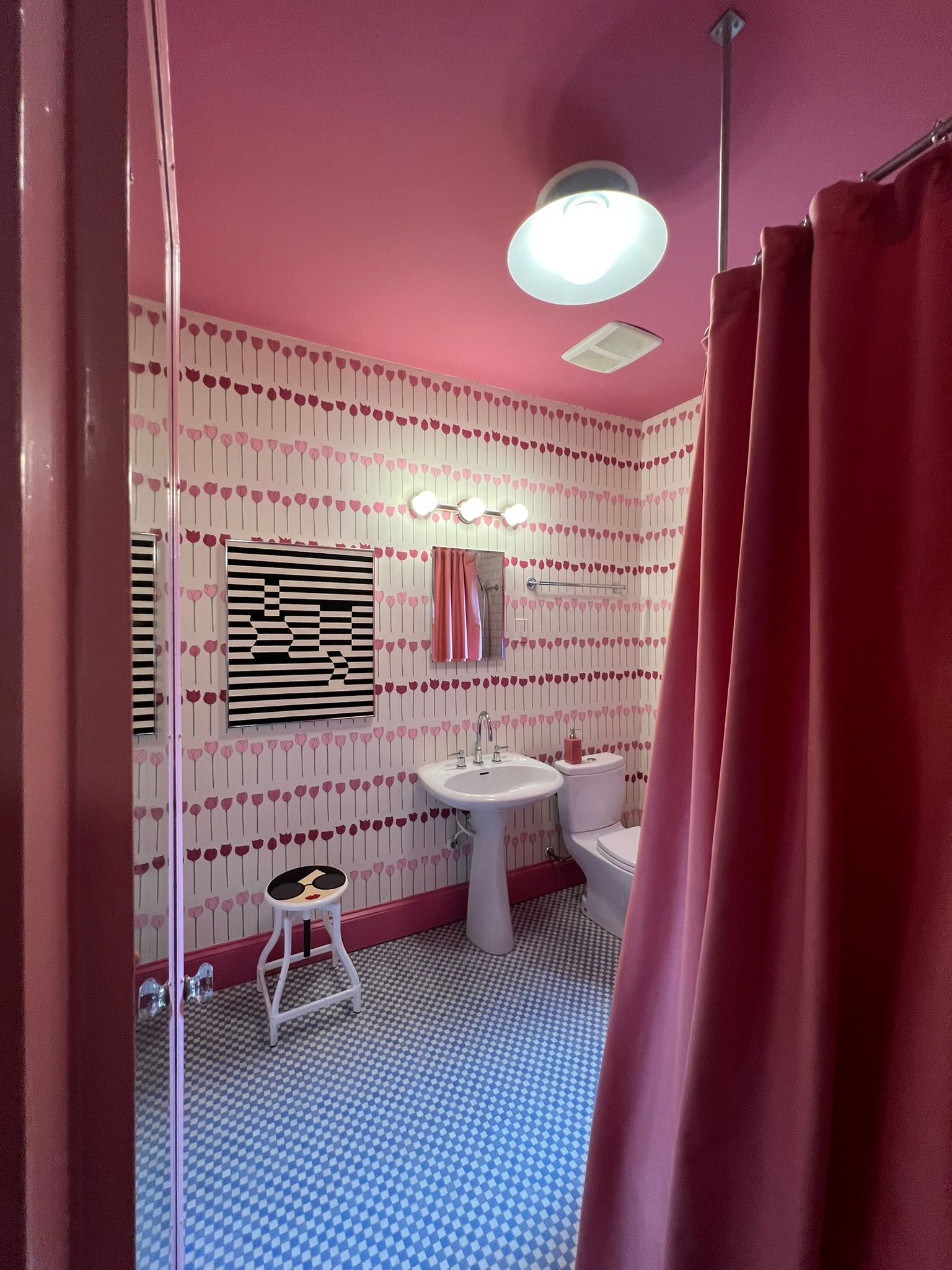 Pink bathroom with patterned tile, red shower curtain, sink, stool, and overhead light.