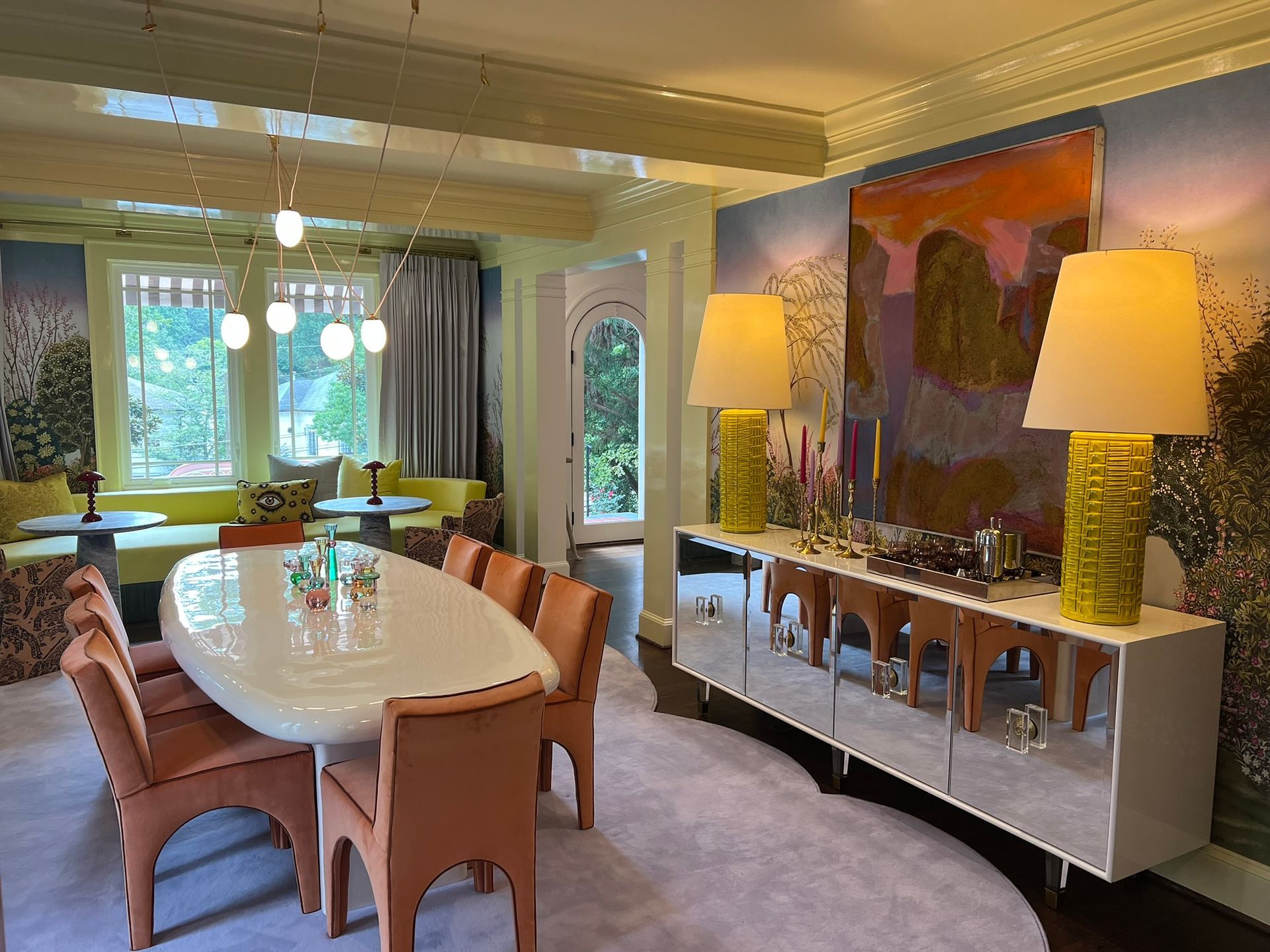 Modern dining room with a long table, wicker chairs, large sideboard, lamps, and garden-view windows