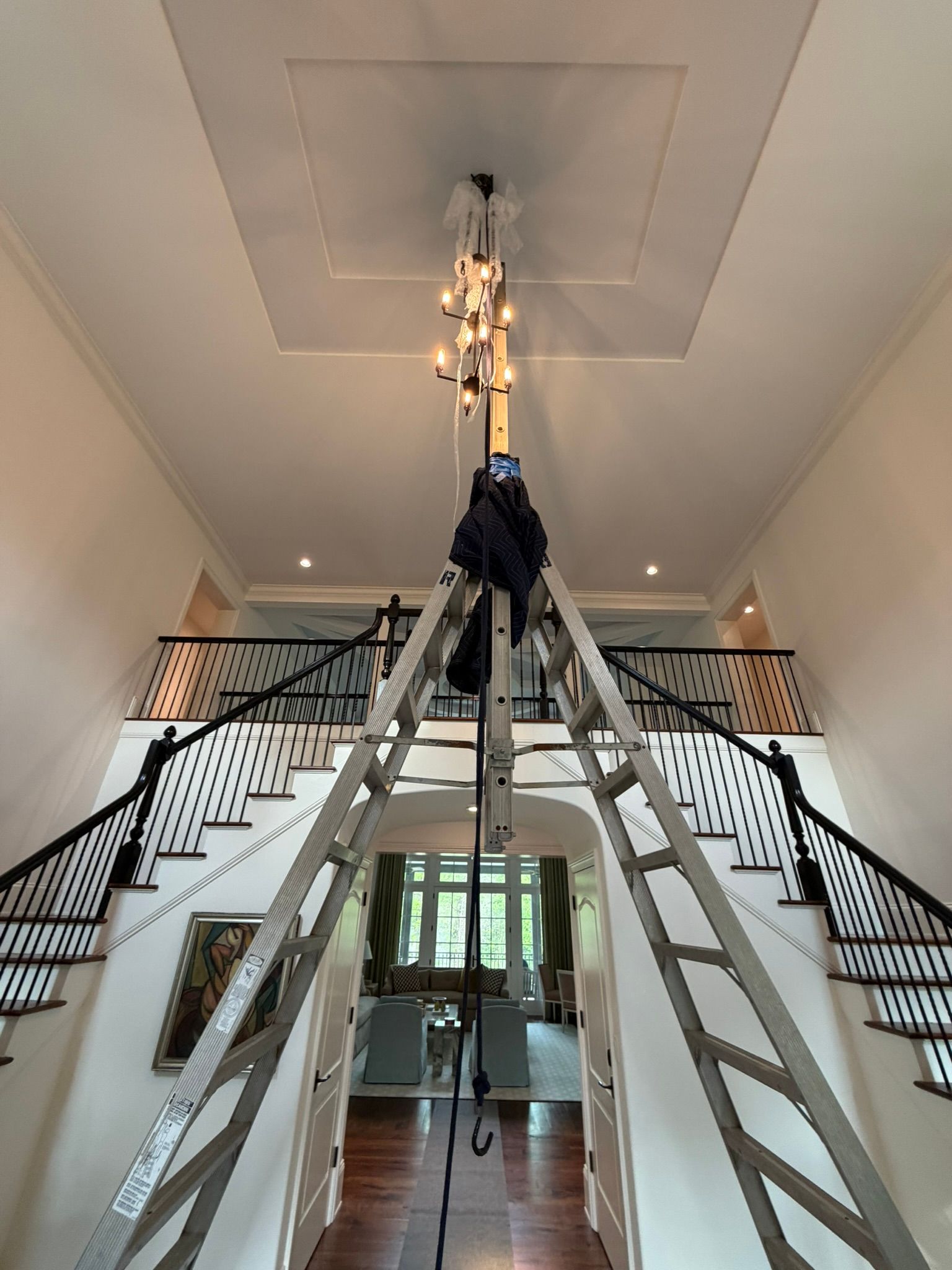 Tall ladder setup in a grand foyer beneath a chandelier, with stairs on both sides.