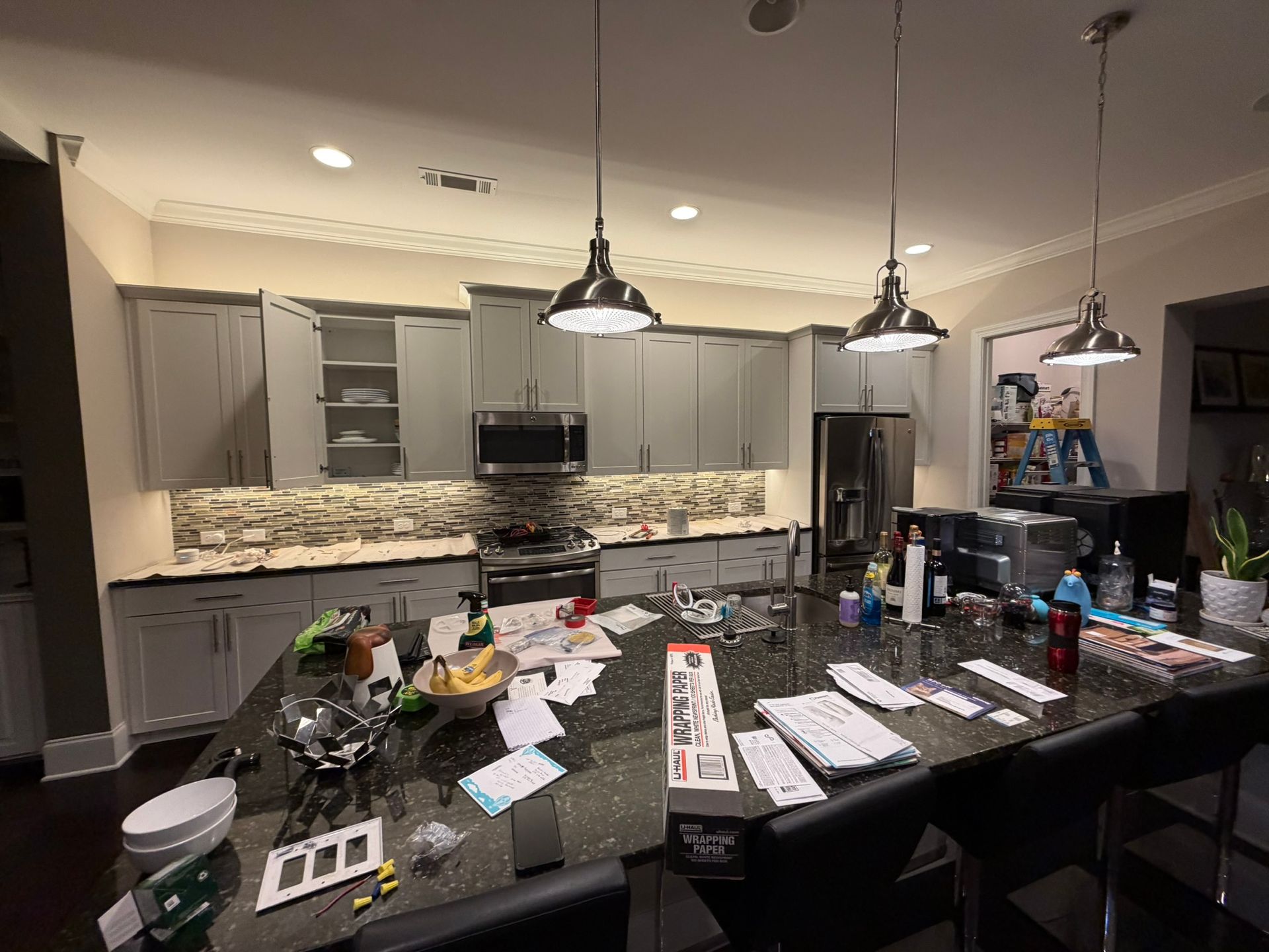Modern kitchen with a large island cluttered with papers, dishes, and bottles under pendant lights.