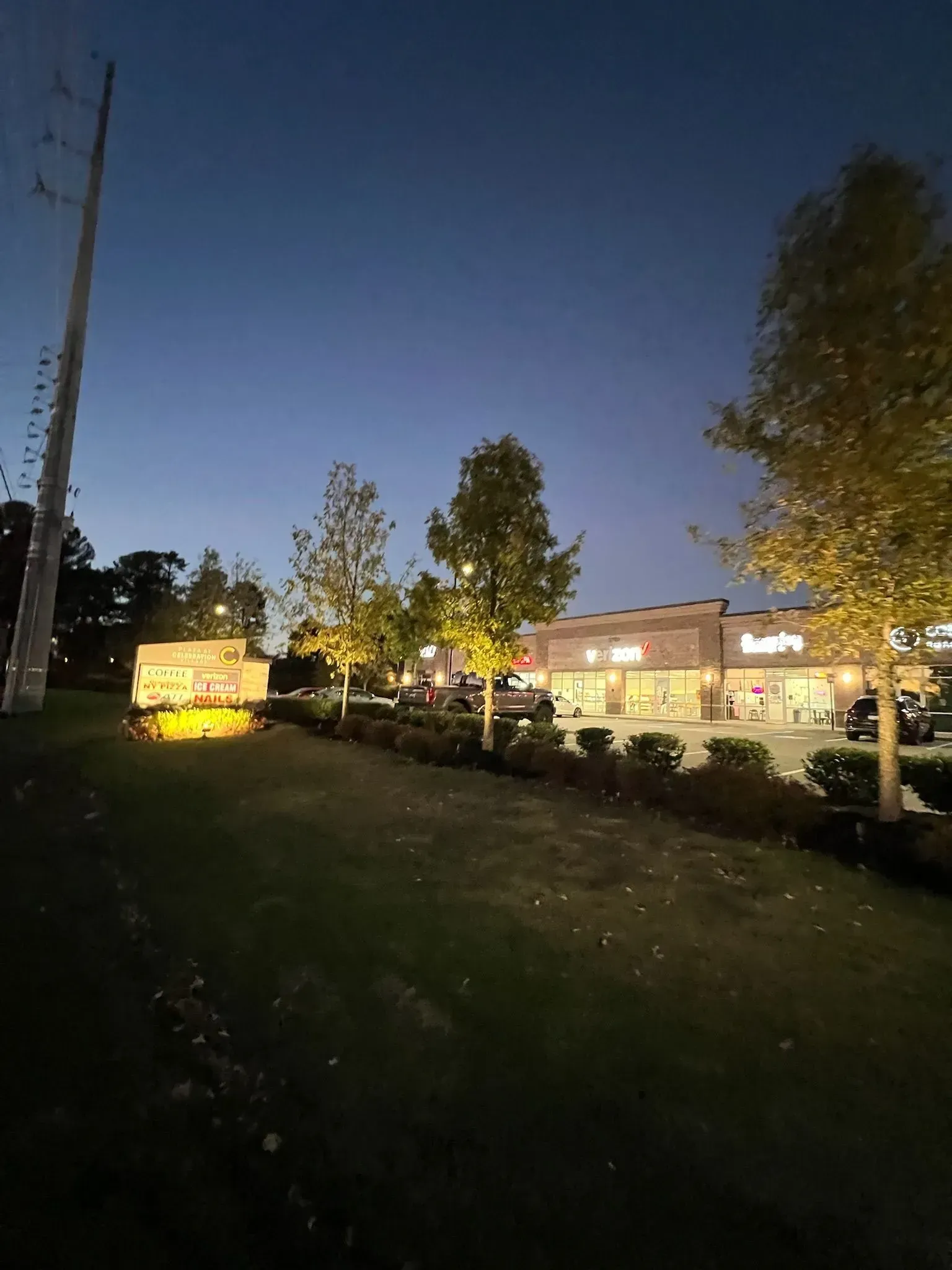 Dusk over a grassy park with lit buildings, trees, and a yellow sign by the road