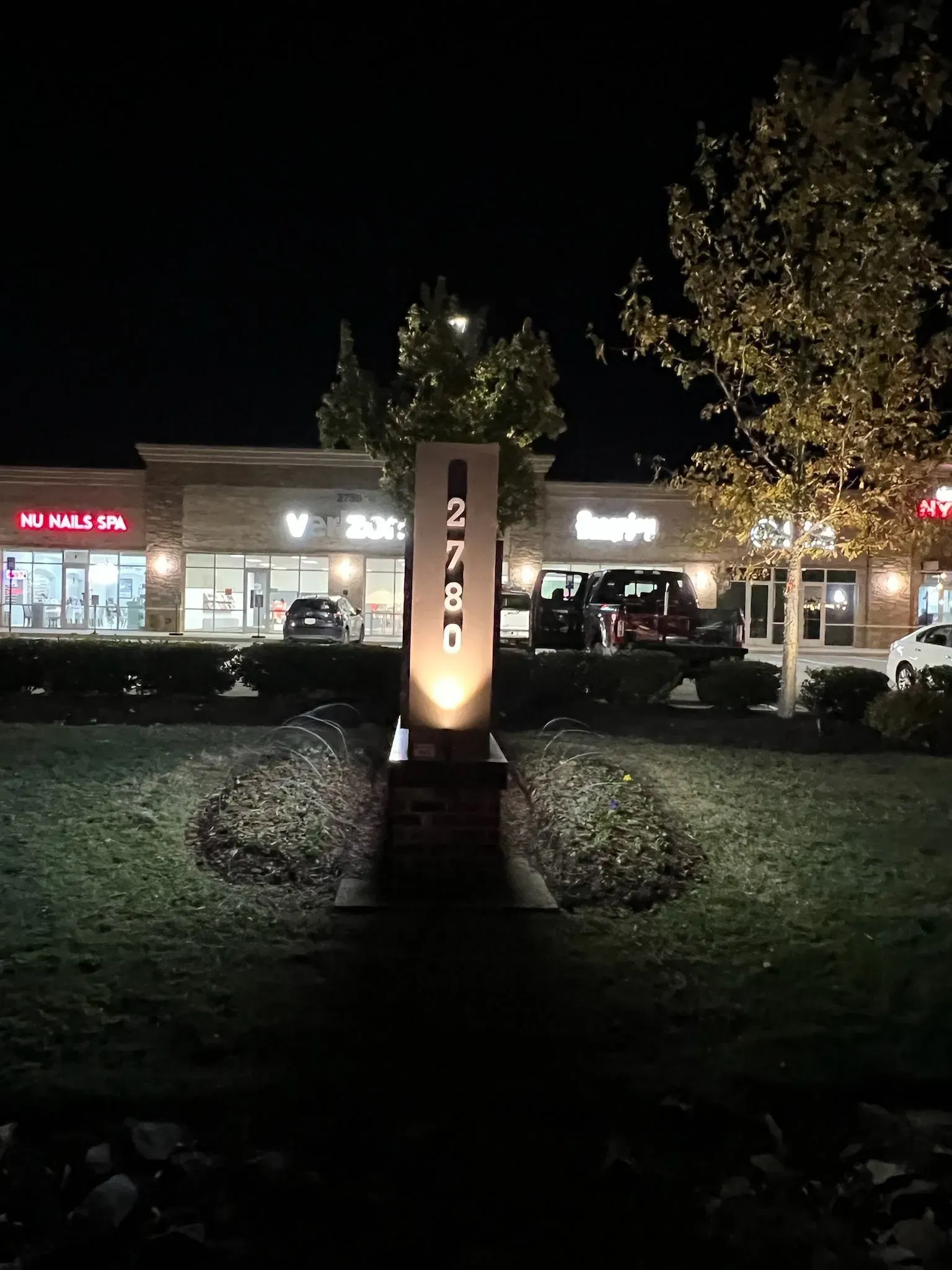 Night view of a lit shopping plaza with trees and a central illuminated sign in the foreground