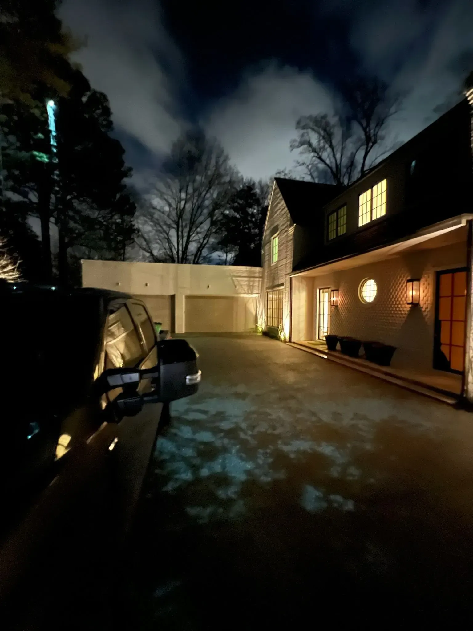 Nighttime driveway beside a lit two-story house, with an open car door and cloudy sky overhead