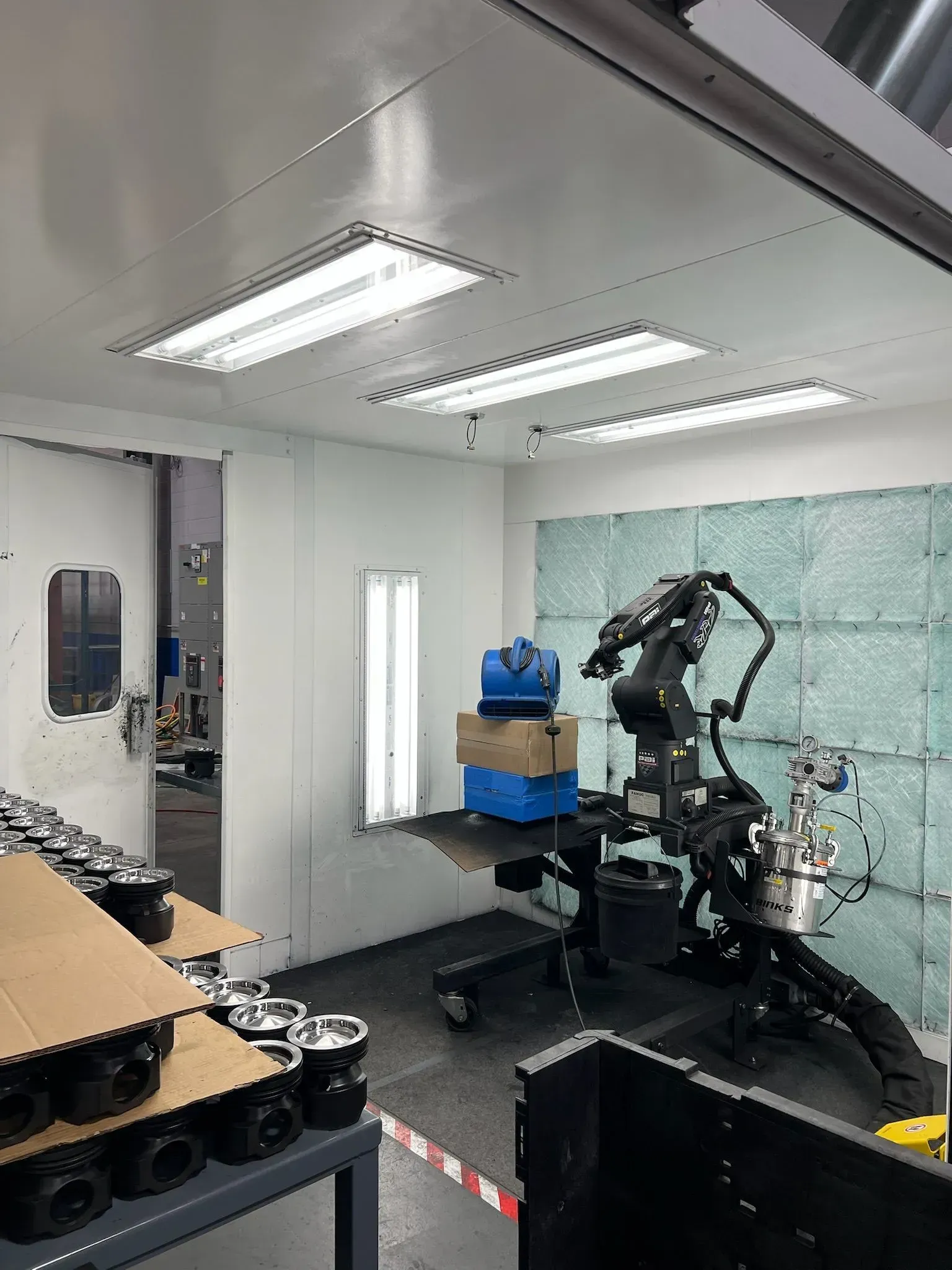 Industrial robot arm in a clean workshop beside a workbench, boxes, and overhead fluorescent lights.