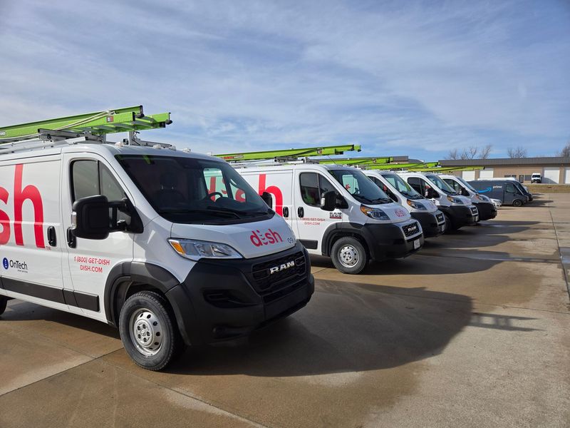 A row of white Dish branded service vans parked in a line on a paved lot under a blue sky.