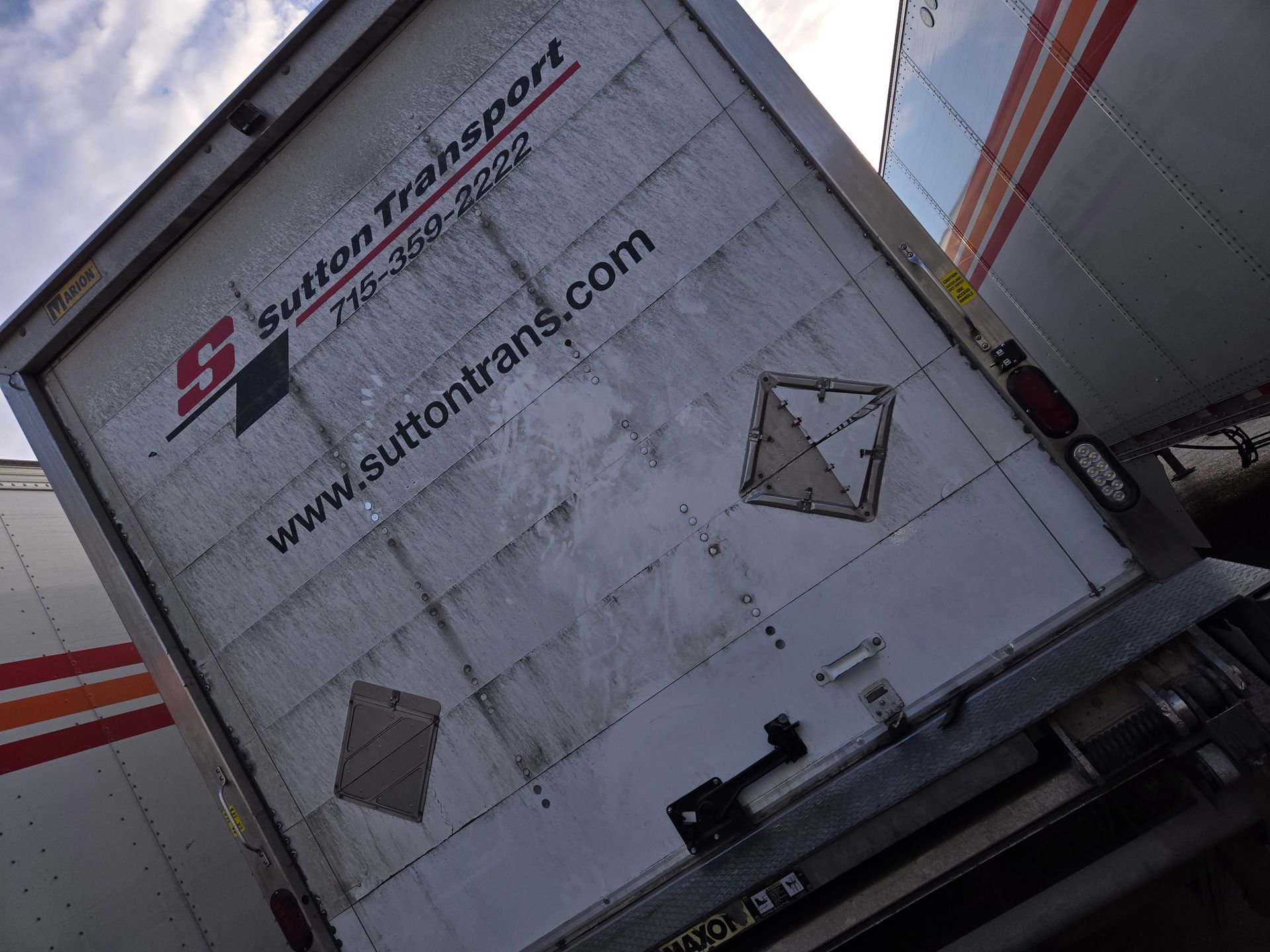 The back of a white Sutton Transport semi-trailer parked outdoors, featuring company branding and a hazard placard.
