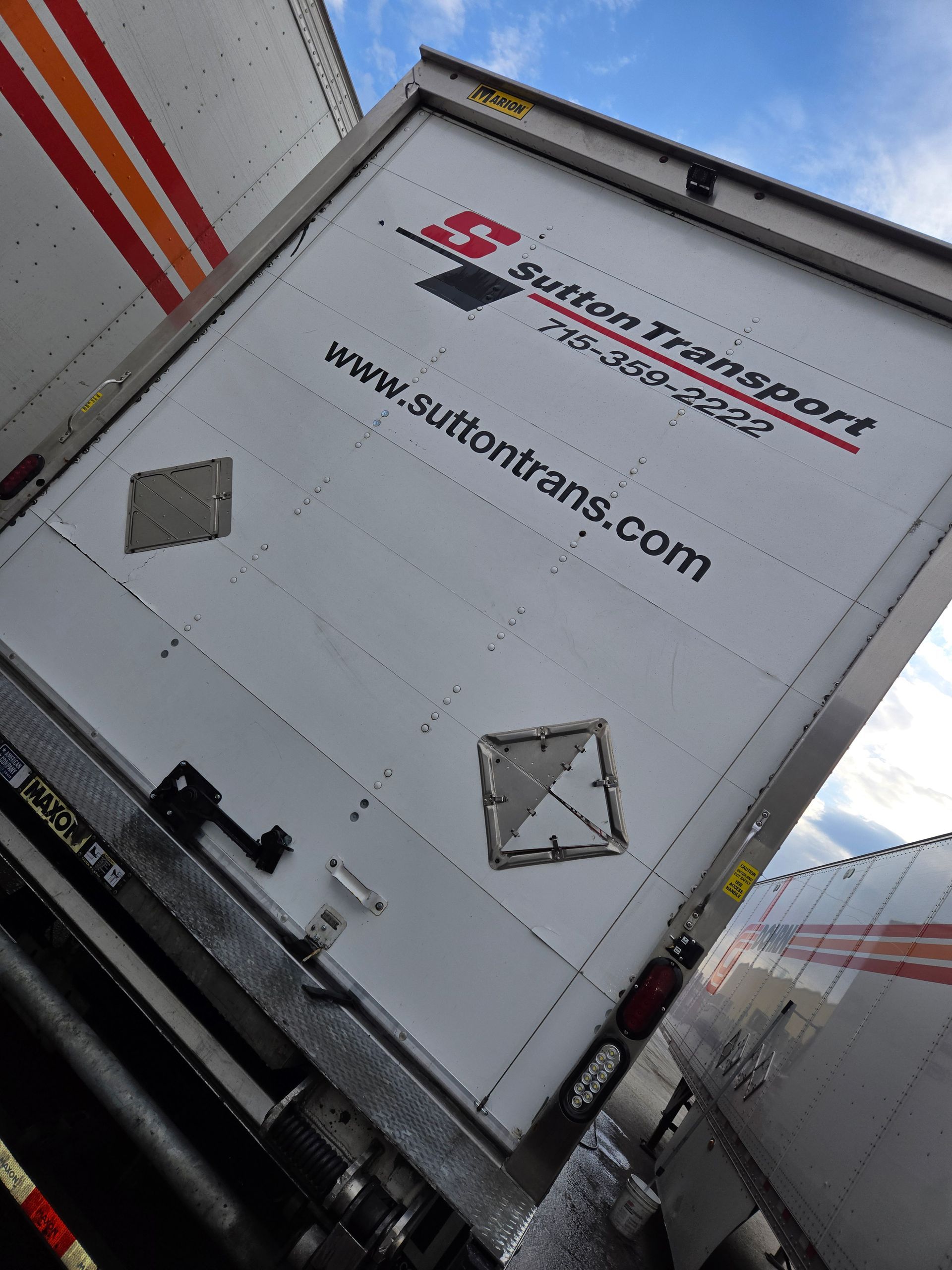 The back of a white Sutton Transport semi-trailer parked outdoors under a blue sky, featuring the company logo and URL.