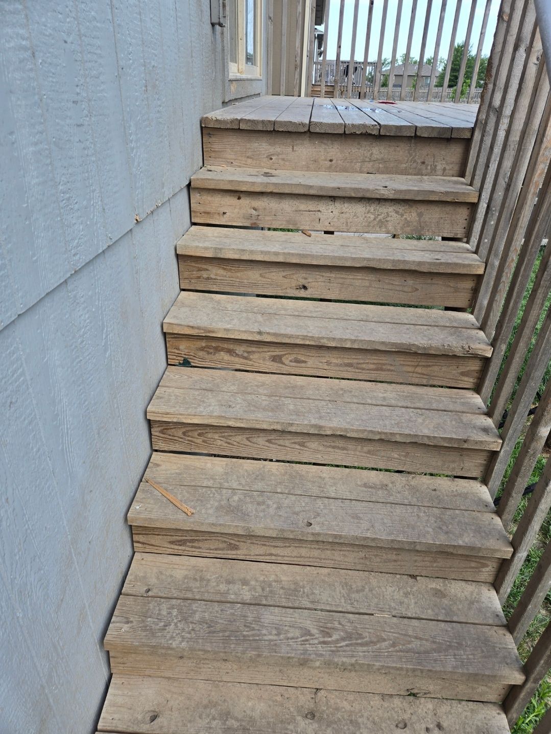 Weathered wooden outdoor stairs with a gray siding wall on the left and a wooden railing on the right.