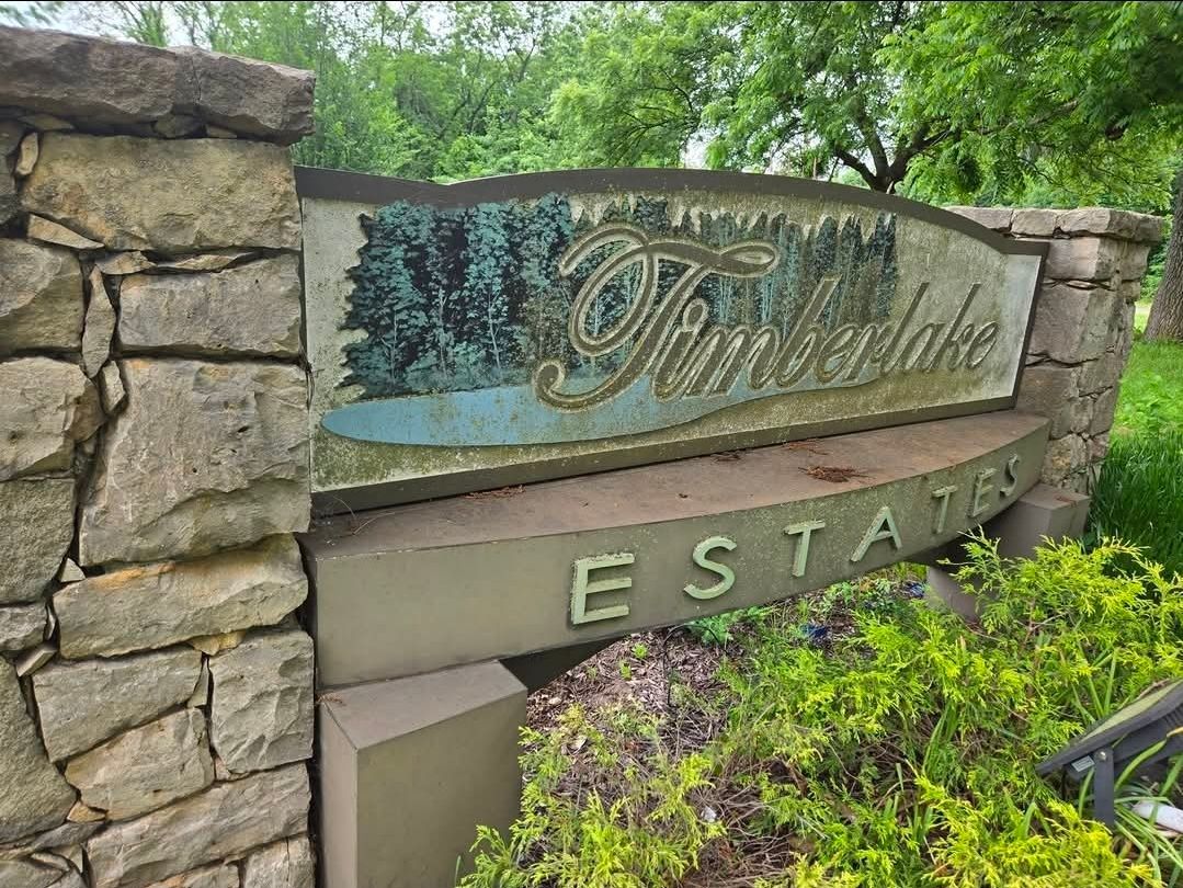 A stone sign for Timberlake Estates featuring cursive text and an illustration of a forest and pond.